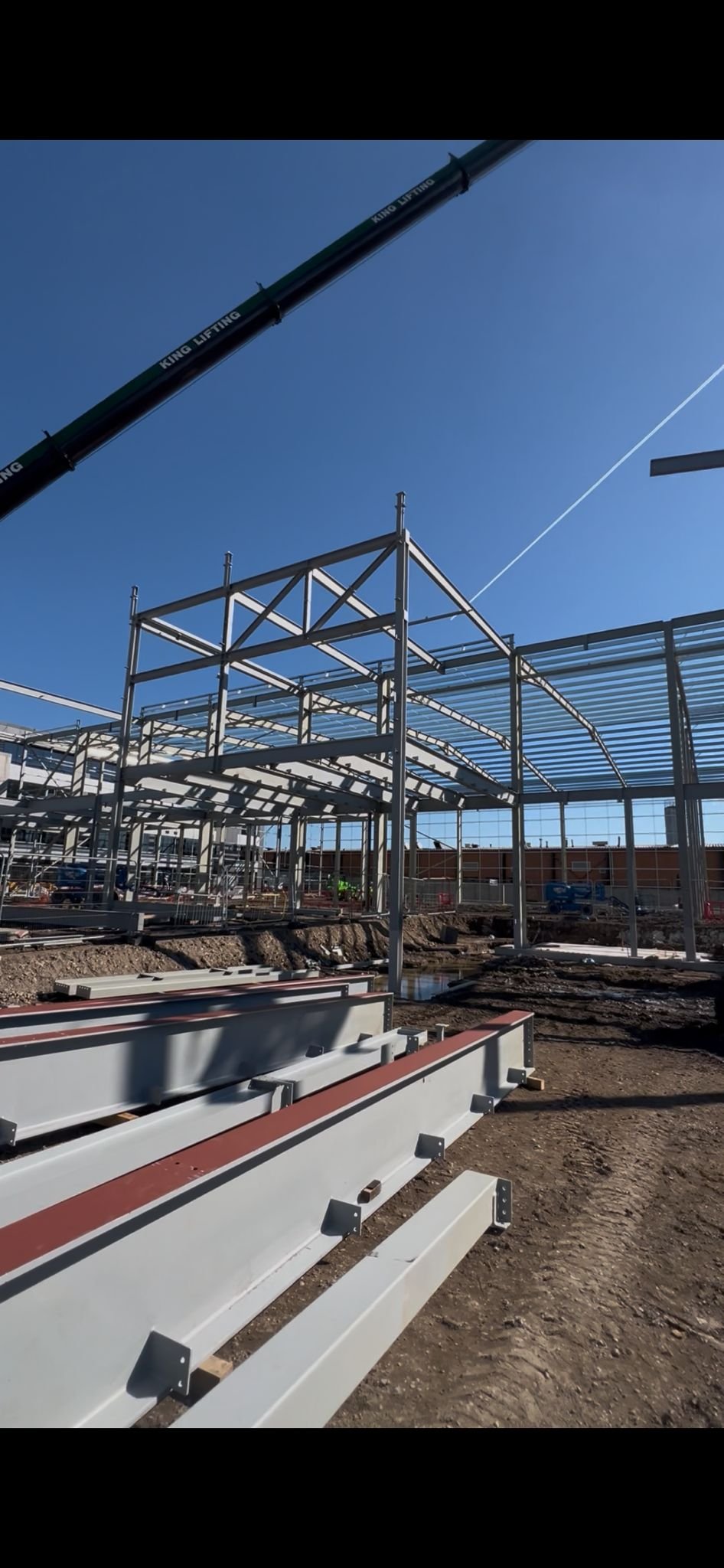 Construction site with a partially built metal structure, construction workers, and equipment. A worker in an orange safety vest stands in the foreground. Gravel on the ground and safety barriers are around the site.