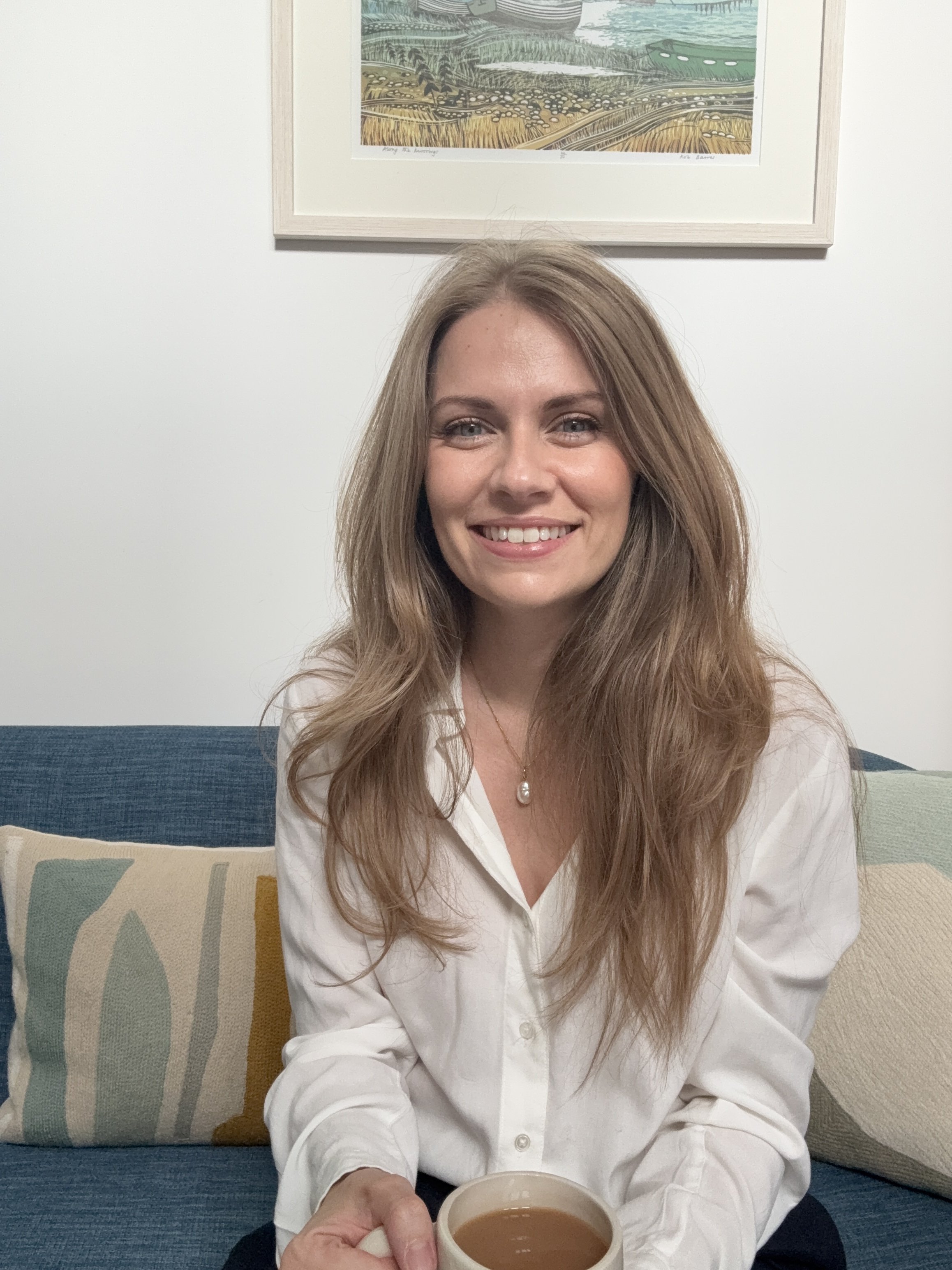 A woman with long, wavy, blonde hair sitting on a colorful couch, smiling, holding a mug of coffee or tea, with a framed artwork above her on a white wall.