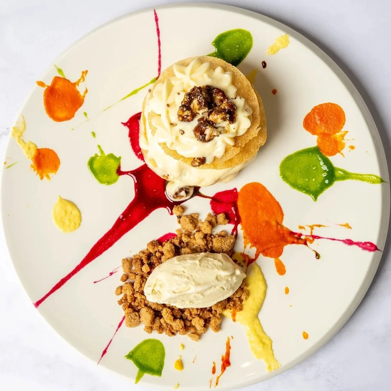 Dessert plate with colorful splashes of red, orange, yellow, and green sauces, featuring a small cake topped with cream and walnuts, and a crumbly piece with a scoop of vanilla ice cream.