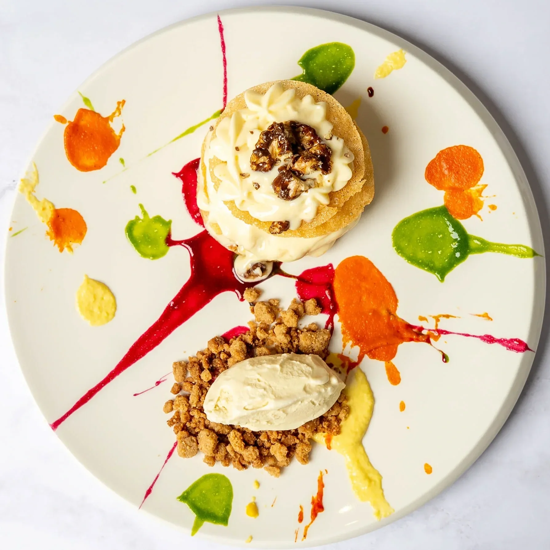 Dessert plate with two ice cream scoops, one topped with whipped cream and nuts, colorful sauce splatters, and crumbled topping, on a white plate.