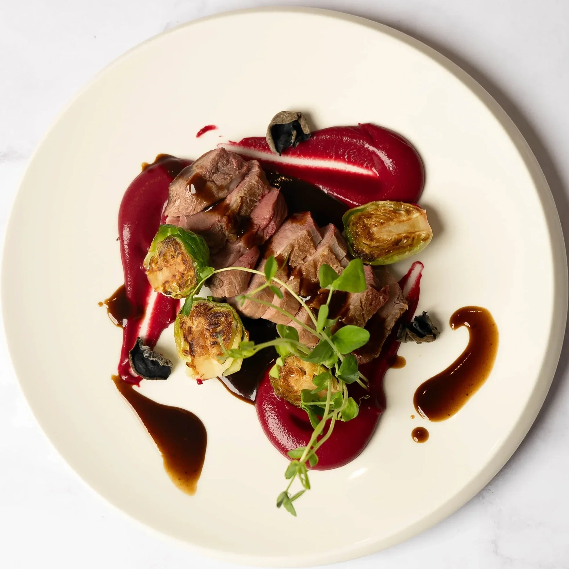 A gourmet dish with sliced beef, roasted Brussels sprouts, red and black sauces, garnished with microgreens on a white plate.