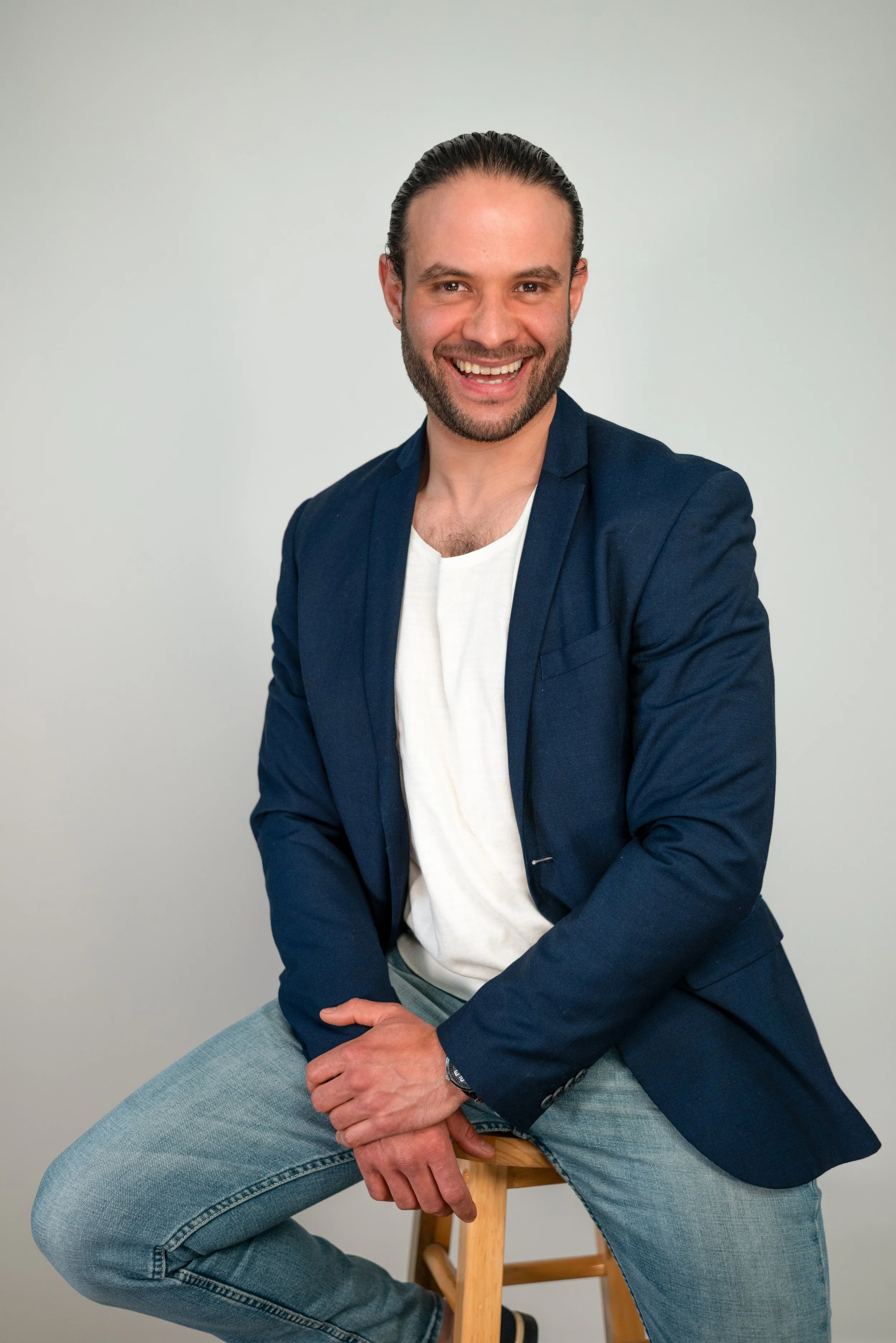 A smiling man in a blue blazer, white T-shirt, and jeans, sitting on a wooden stool against a plain gray background.