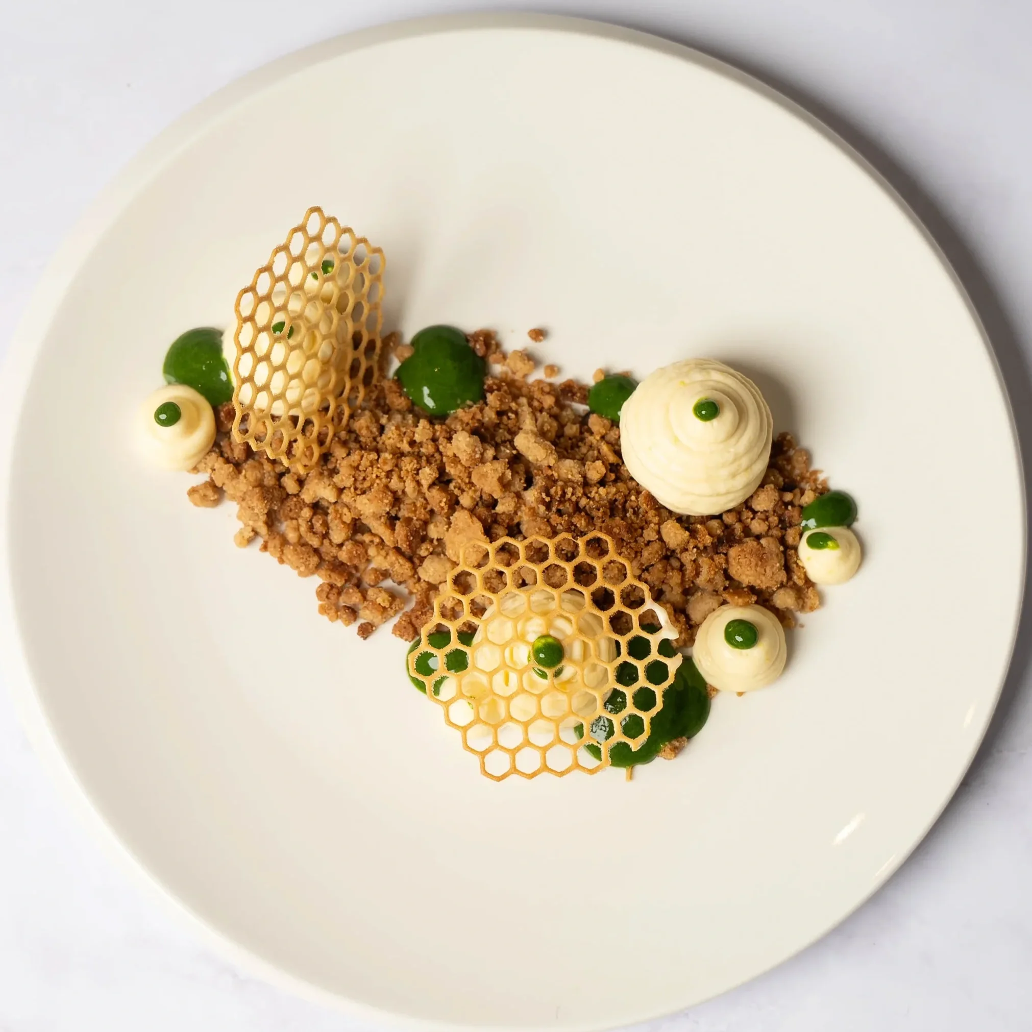 A plated gourmet dessert with crumble, green dots, white cream swirls, and honeycomb-like caramel decorations on a white plate.