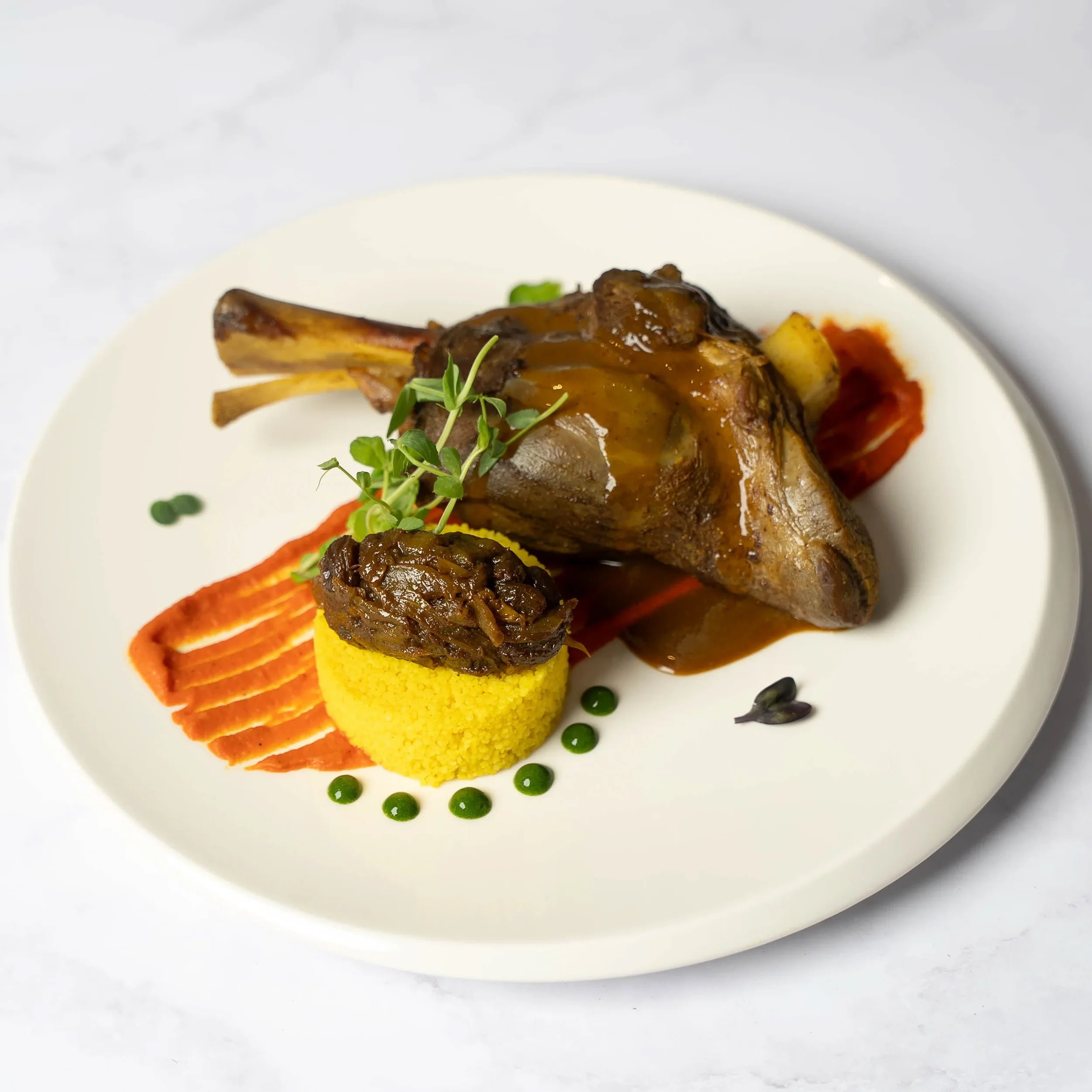 Plate with cooked lamb chop topped with brown sauce, garnished with microgreens, served with yellow polenta and vegetables, decorative sauce dots on white plate.