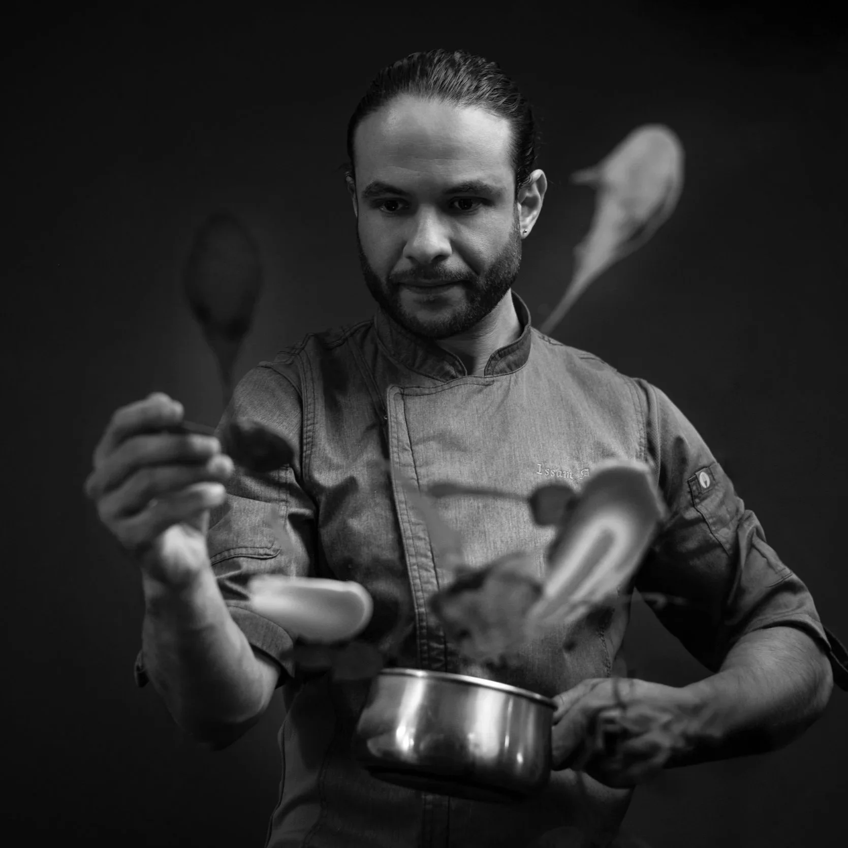 A man dressed in a chef's coat tossing ingredients into a pot. He has a focused expression and is holding a spoon and some vegetables. The image is black and white.