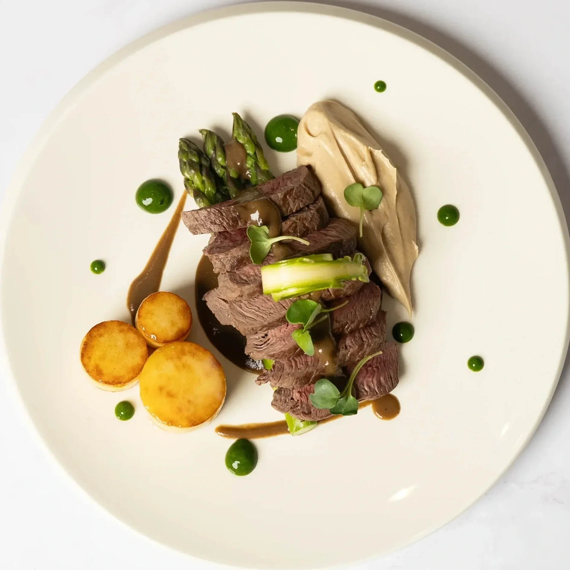 Gourmet plate with slices of beef, asparagus, roasted potatoes, a smear of creamy sauce, and green herb droplets.