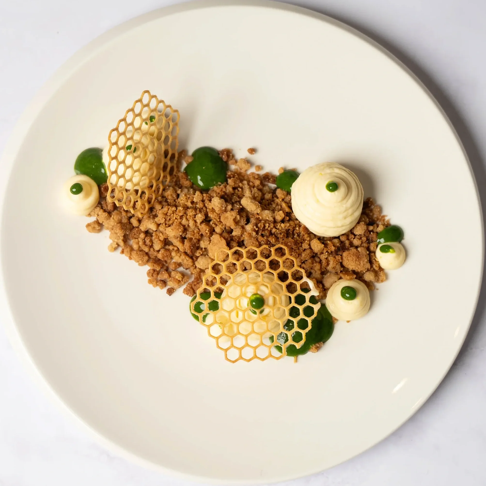Modern plated dessert with crumbly base, white cream dollops topped with green sauce, and decorative honeycomb-like sugar structures on a white plate.