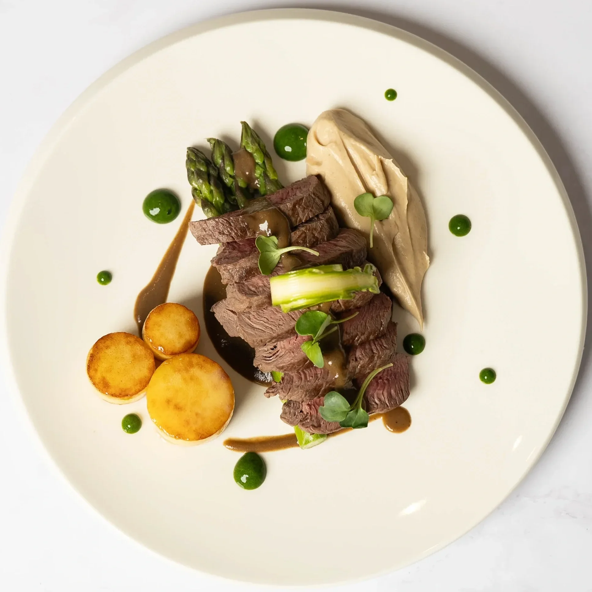 Gourmet meat dish with sliced steak, asparagus, roasted potatoes, pâte, microgreens, and sauce drizzles on a white plate.