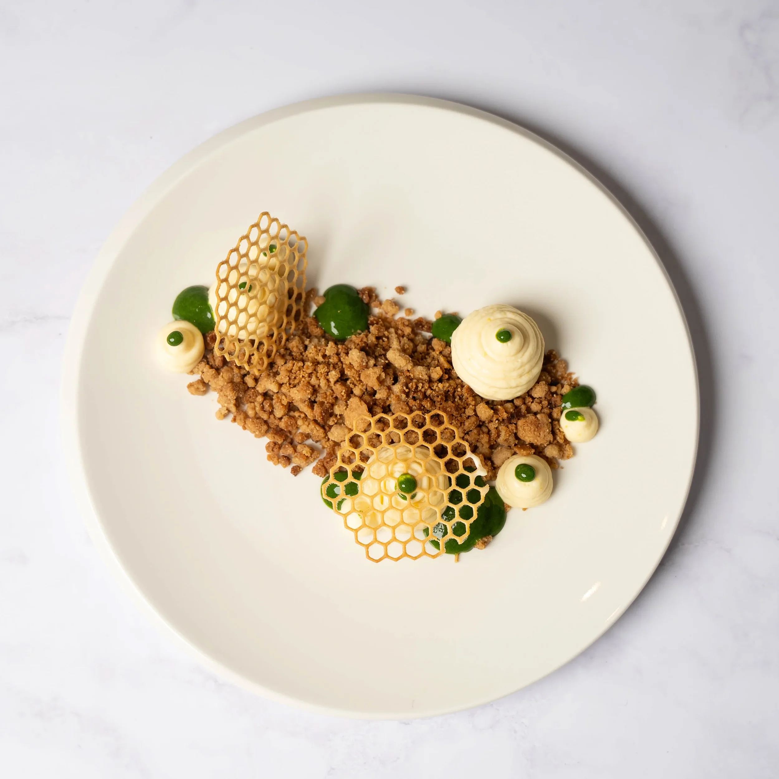 Elegant plated dessert with crumbled cookie base, white chocolate dollops topped with green accents, and decorative honeycomb-like sugar structures.