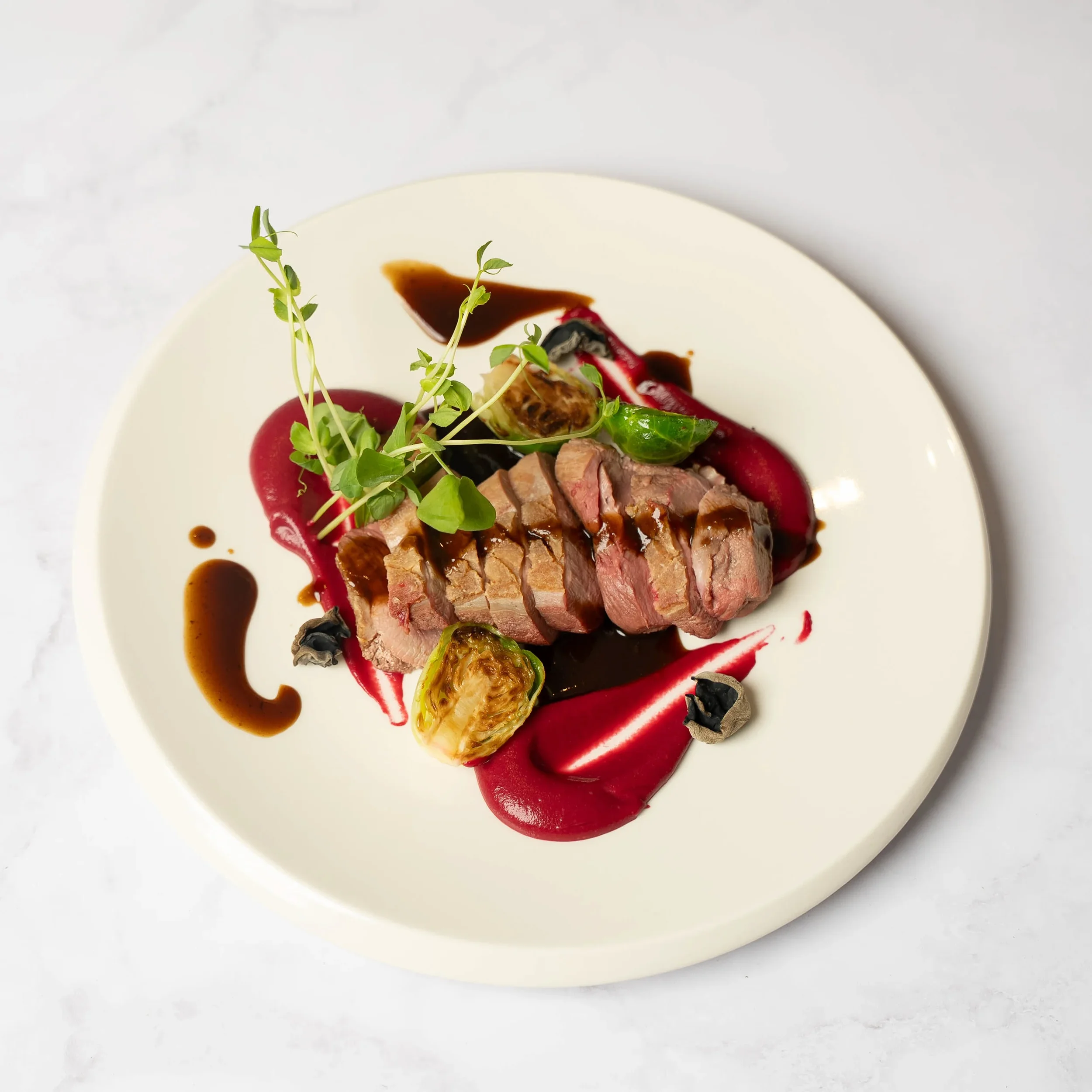 Sliced cooked steak on a white plate with red and black sauces, garnished with microgreens, roasted Brussels sprouts, and black and mushroom slices.