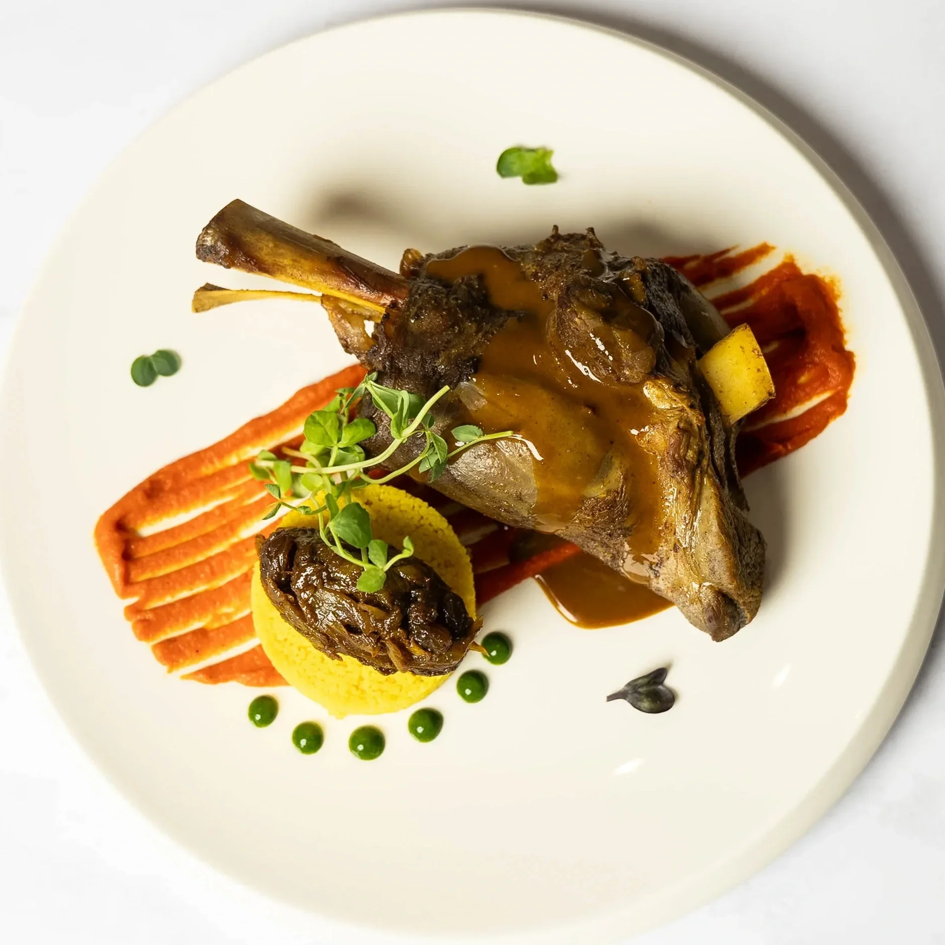 Gourmet plated lamb chop with sauce, garnished with microgreens, served with a side of mashed sweet potatoes, grilled vegetable, and sauces on a white plate.