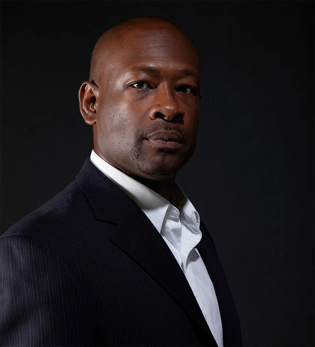 A man with dark skin, bald head, and wearing a black suit and white shirt against a black background.