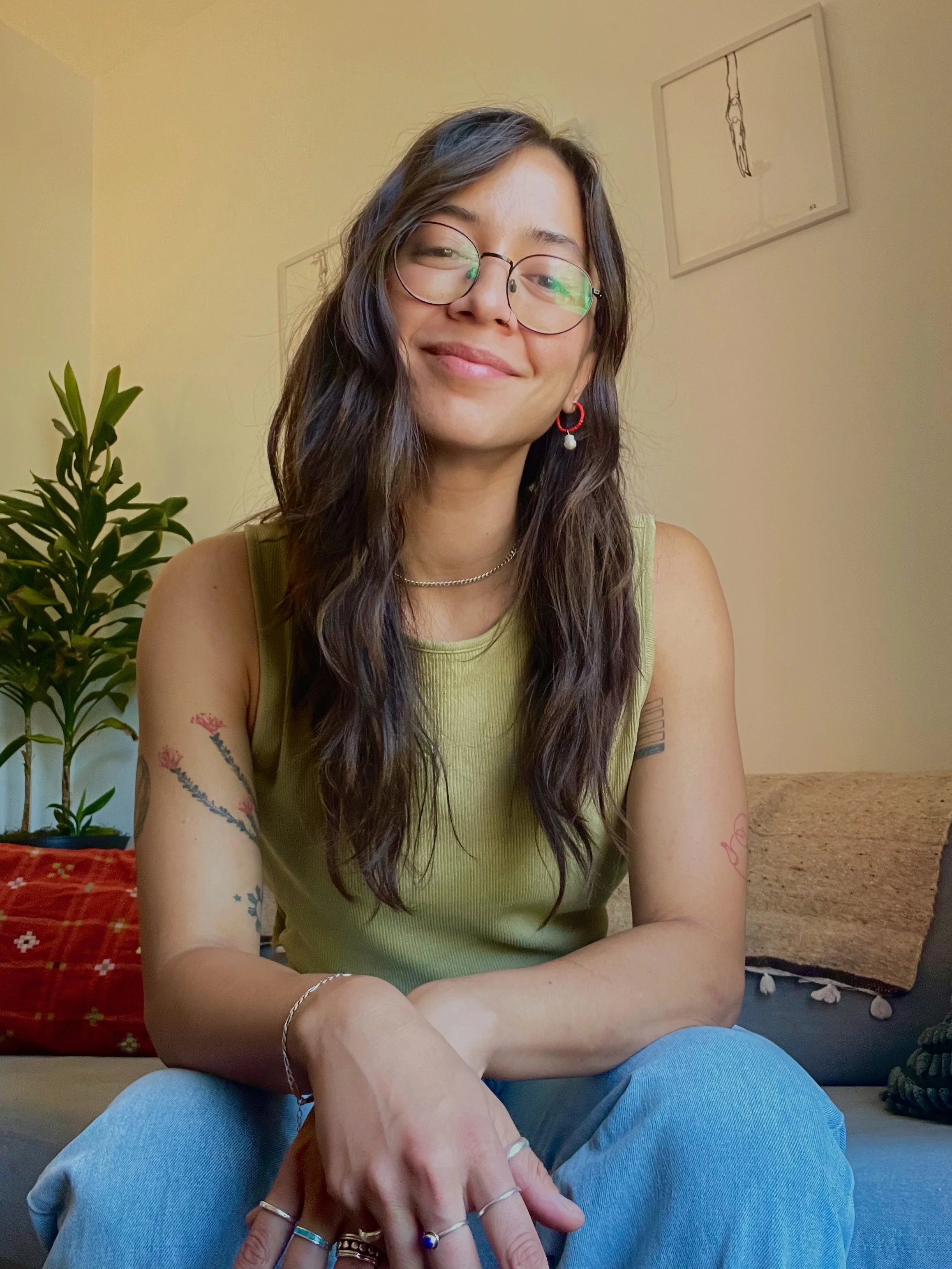 Smiling woman with long wavy hair, glasses, earrings, and tattoos sitting on a couch in a cozy room with plants and framed artwork in the background.