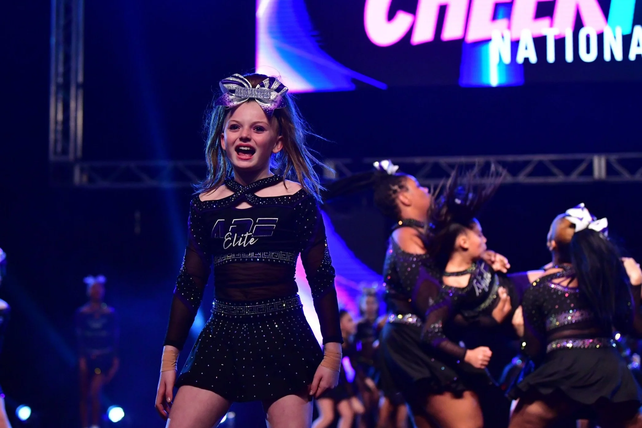 A young girl in a black cheerleading outfit with rhinestones stands on stage, smiling and cheering at a cheerleading competition, with other cheerleaders performing behind her.