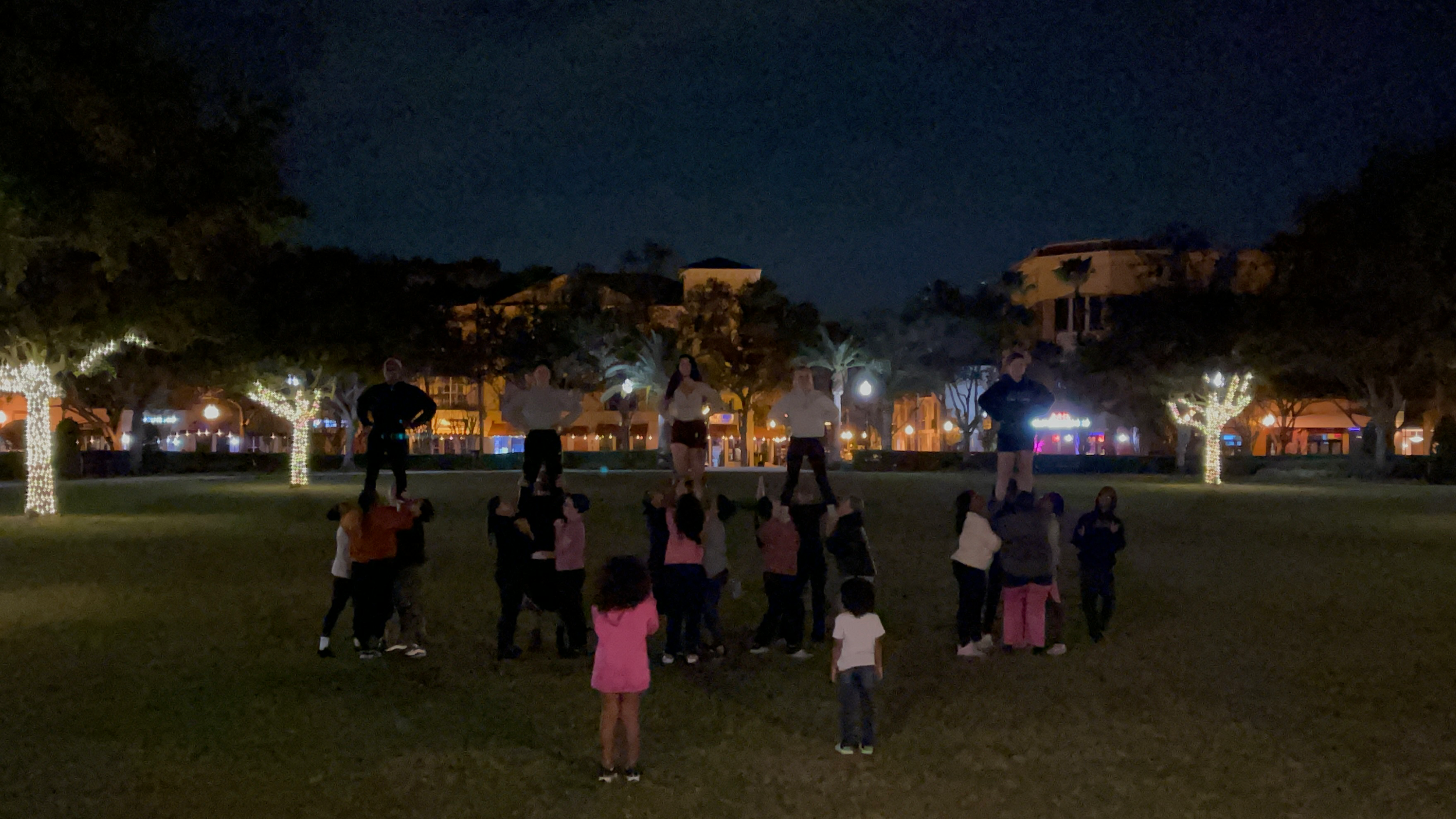 Night scene with people gathered outside, some standing on elevated platforms, and trees decorated with string lights in a park-like setting.