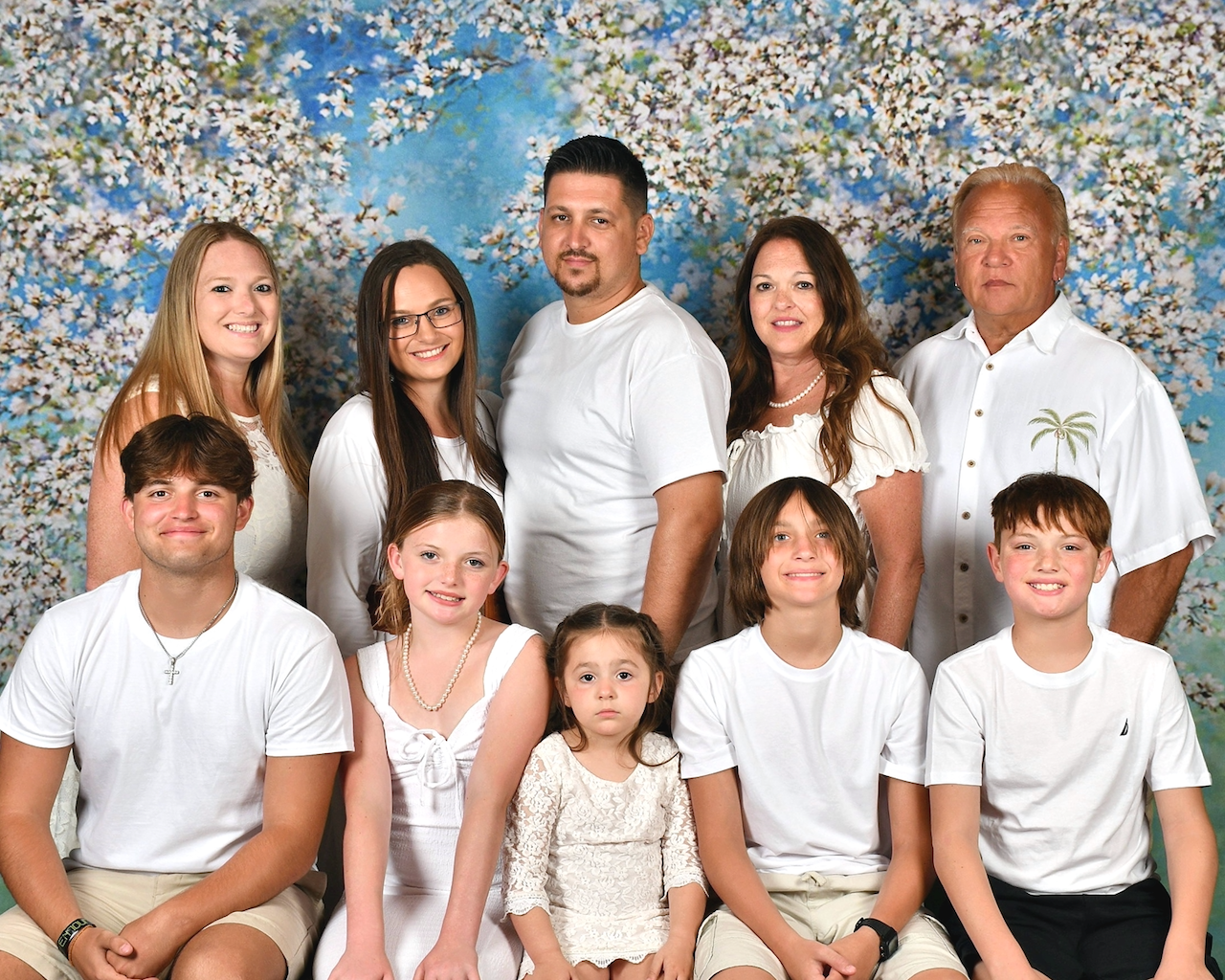 Family portrait of ten people, including adults and children, dressed in white clothing, smiling, and standing in front of a backdrop with a floral and blue sky pattern.