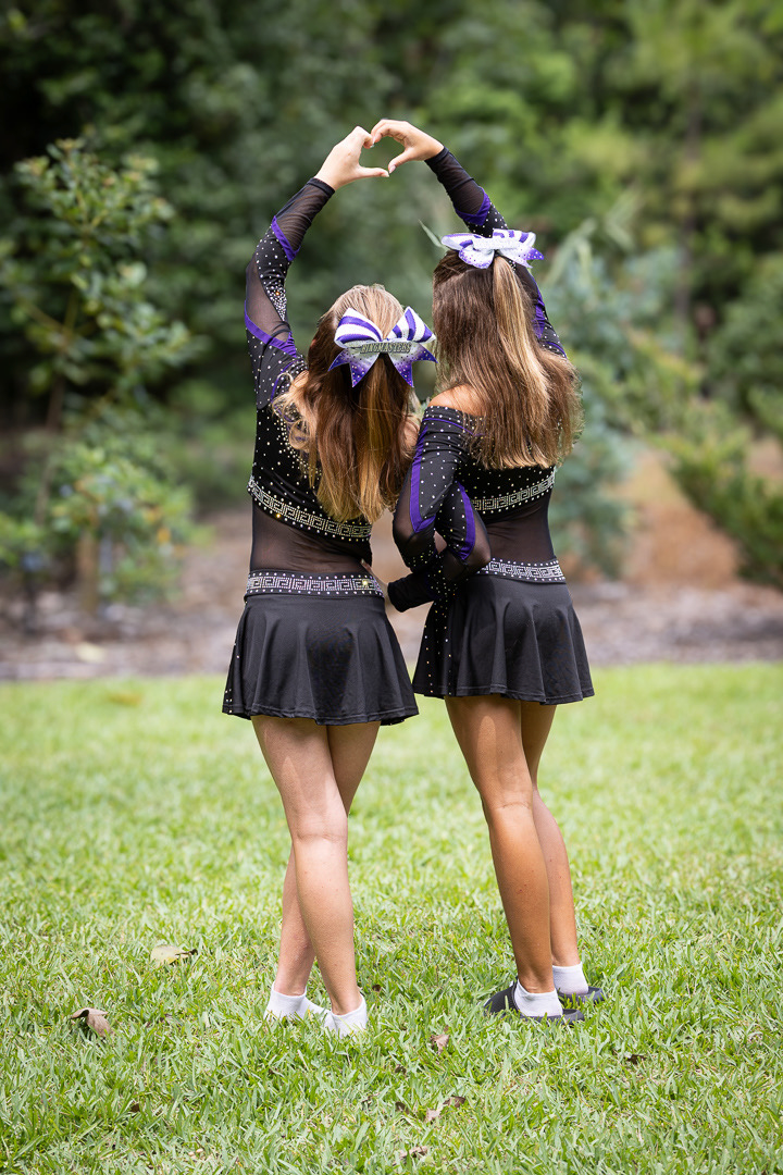 Two cheerleaders dressed in black and purple uniforms standing outside on green grass, facing away and forming a heart shape with their arms.