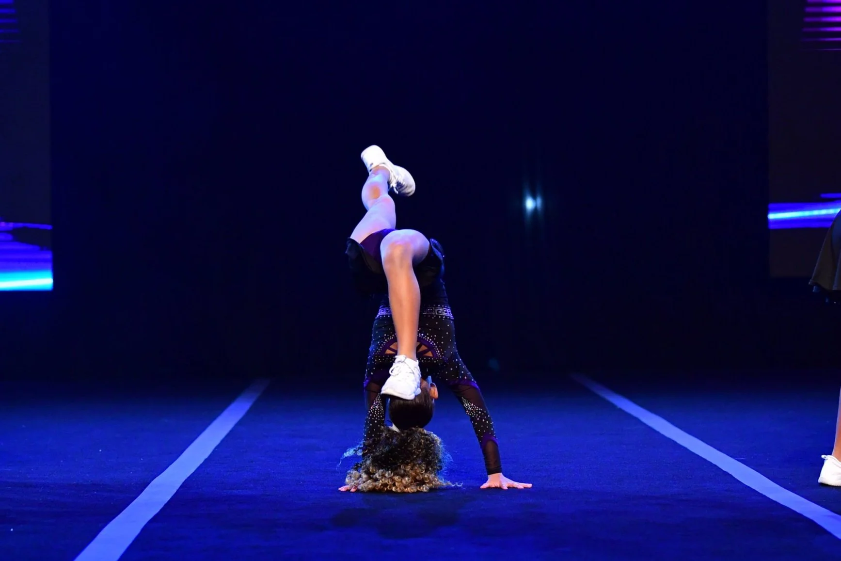 Two cheerleaders performing a partner stunt. One cheerleader is upside down with hands on the floor, supporting the other who is balancing on her shoulders with one leg bent and the other extended upward, wearing a black outfit with sparkling designs.