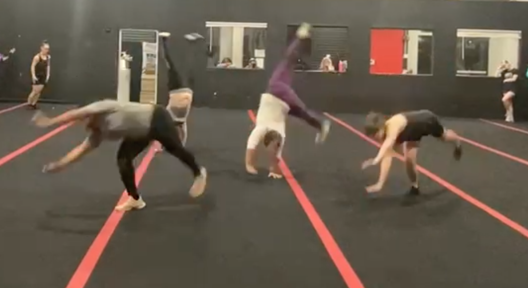 People practicing handstands and acrobatics on a black gym floor with red lines, in a gym with black walls and mirrors.