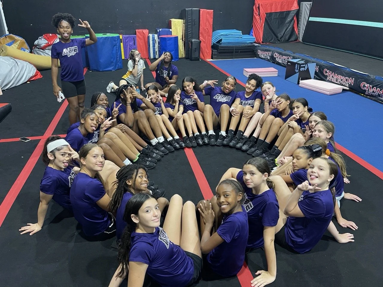 A group of young girls in matching purple athletic uniforms sitting in a circle on a gymnastics gym floor, posing happily for the camera.