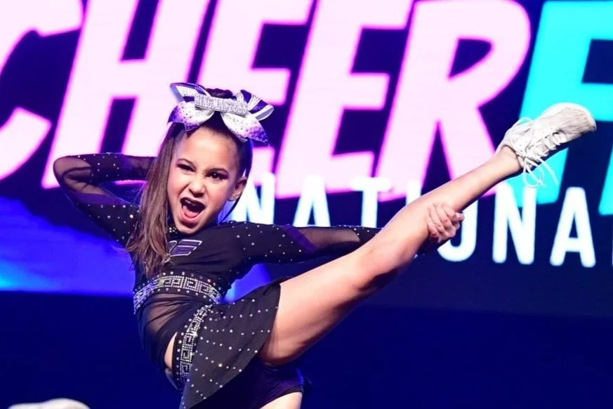 Young girl performing a high kick on stage, wearing a black costume with rhinestones and a large bow in her hair, with a colorful stage background.