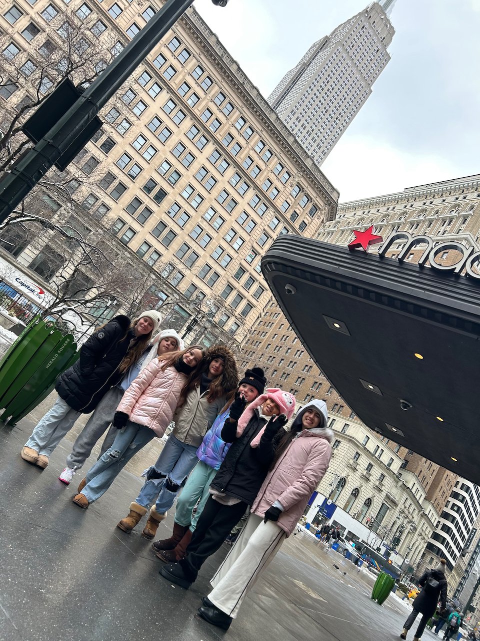 A group of eight young women dressed warmly standing on a city sidewalk in front of a theater marquee in a tall urban area with skyscrapers and historic buildings.