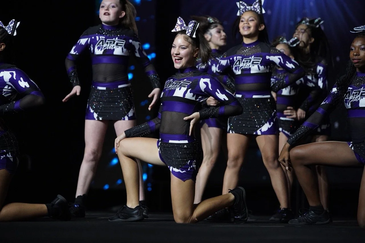 Young female cheerleaders in purple and black uniforms performing on stage, one kneeling with a smile, others standing with hands on hips, all wearing large bows in their hair.