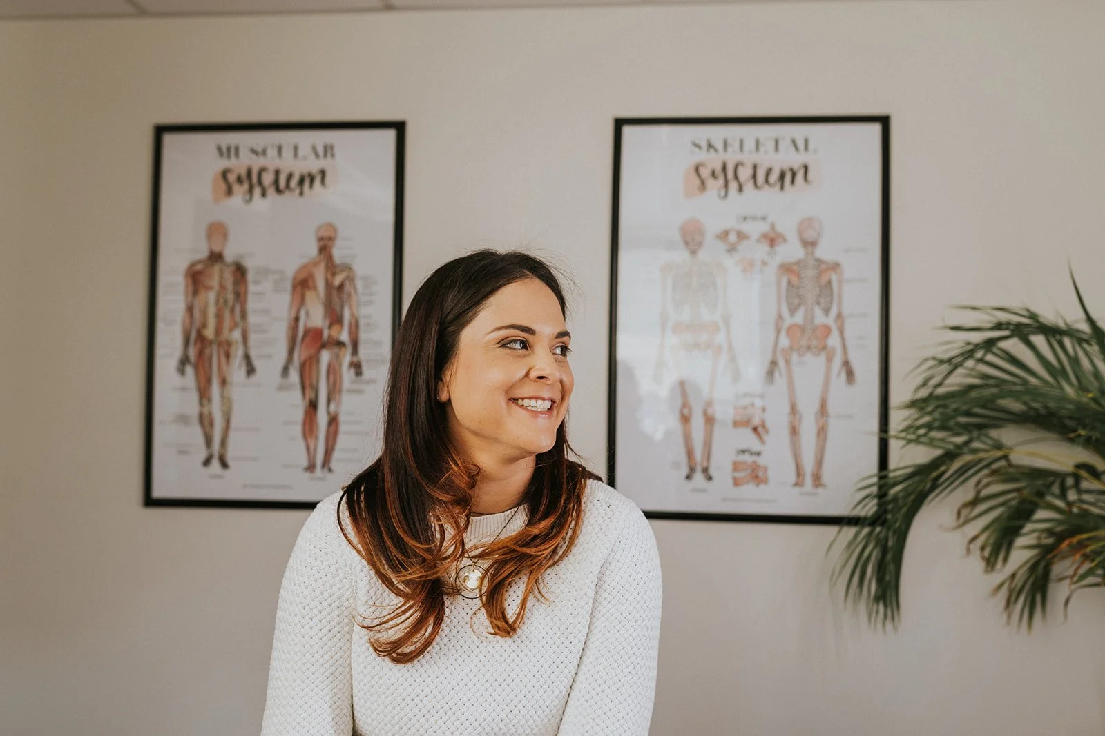 A woman with long brown hair smiling, sitting in front of anatomical charts of muscles and skeletons on a wall, with a green plant partially visible on the right.