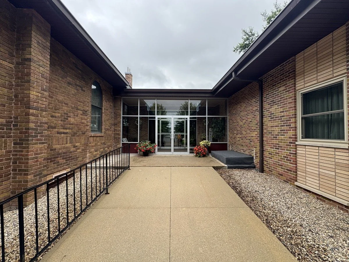 View of building entrance with glass doors, flower pots, brick walls, rain on the ground, overcast sky.