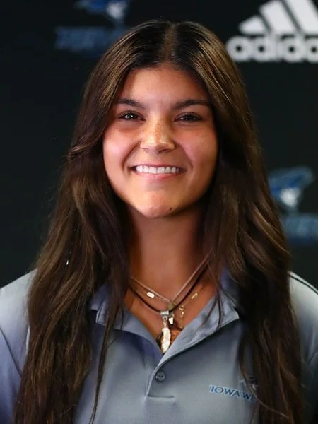 Young woman with long brown hair, smiling, wearing a gray sports shirt and layered necklaces, standing in front of a dark background with logos.