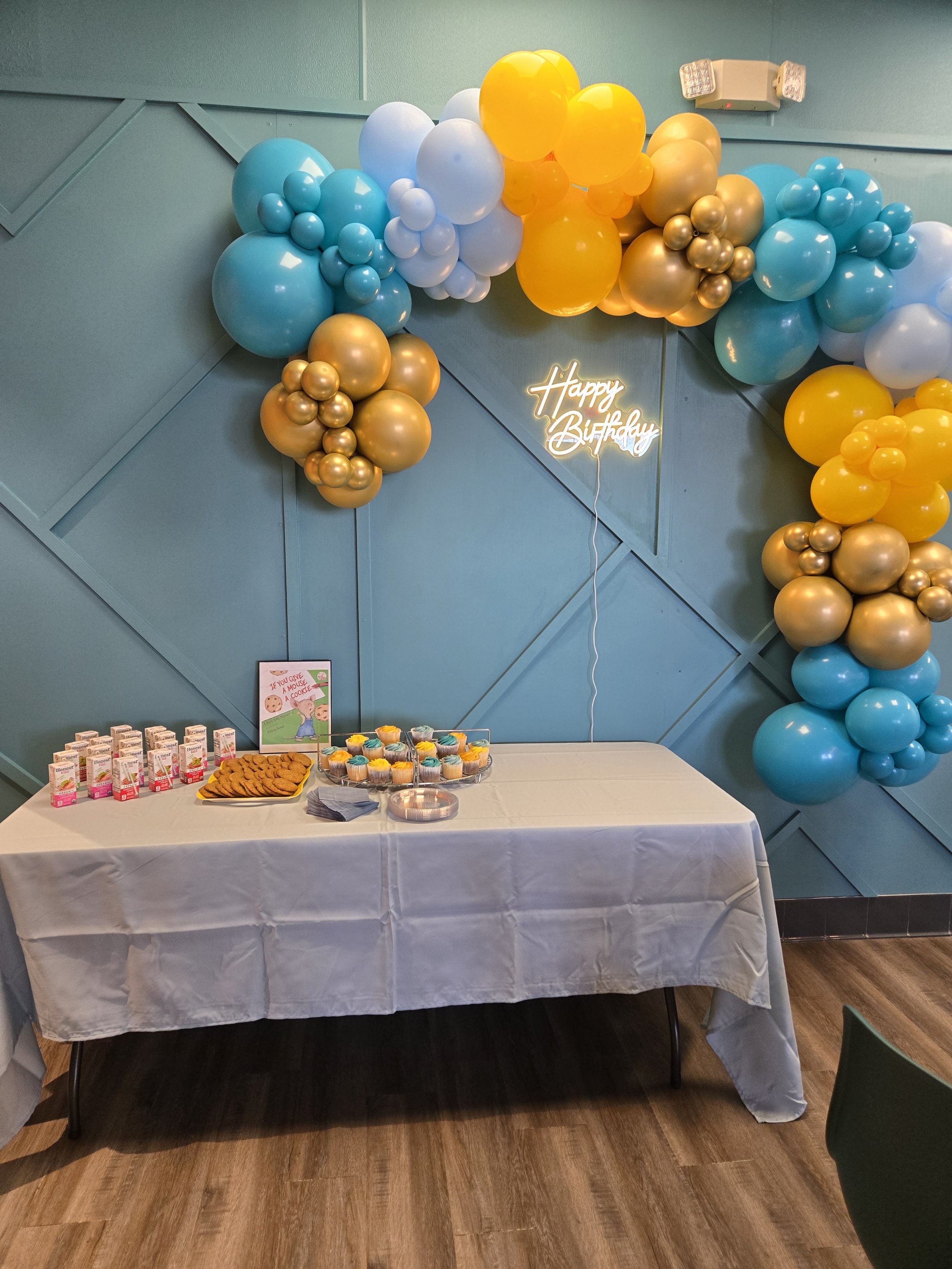 Colorful balloon arch with pink, yellow, green, and orange balloons, decorated with flowers, placed at the entrance of a brick building.