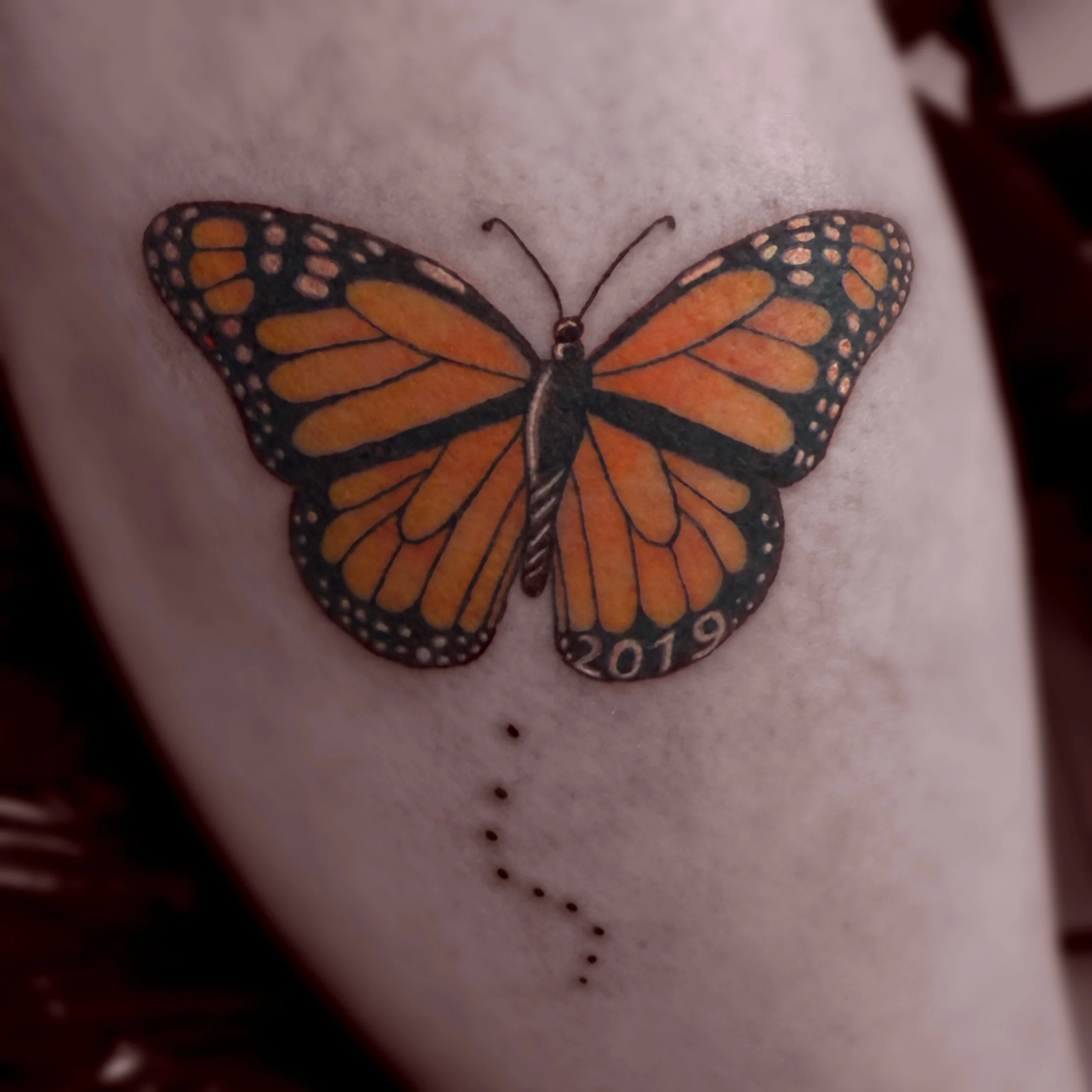 Close-up of a butterfly tattoo on skin, with the year 2019 and a dotted line beneath it.