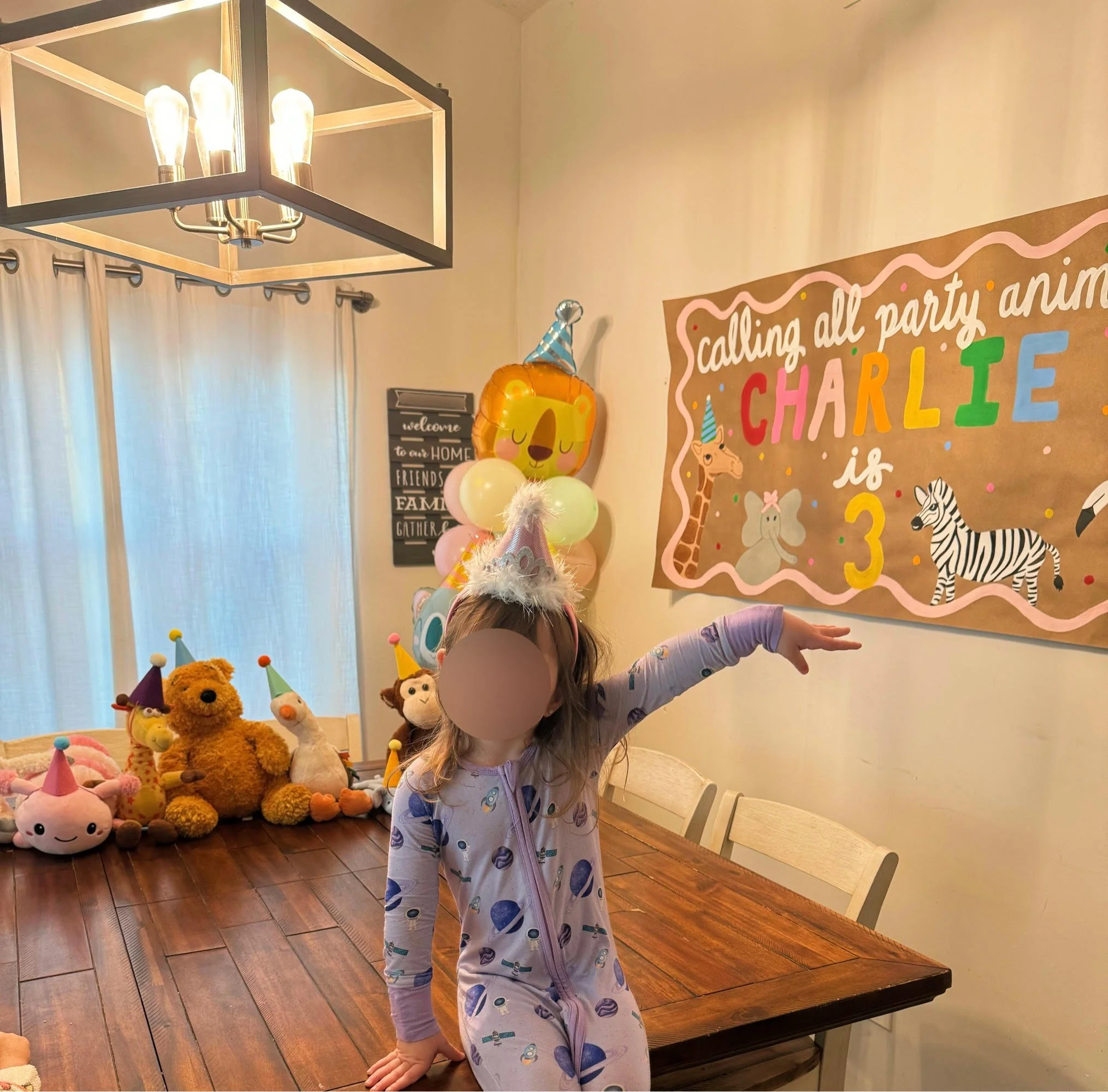 Toddler sitting on table surrounded by birthday decorations such as banner, stuffed animals with party hats, and a balloon column