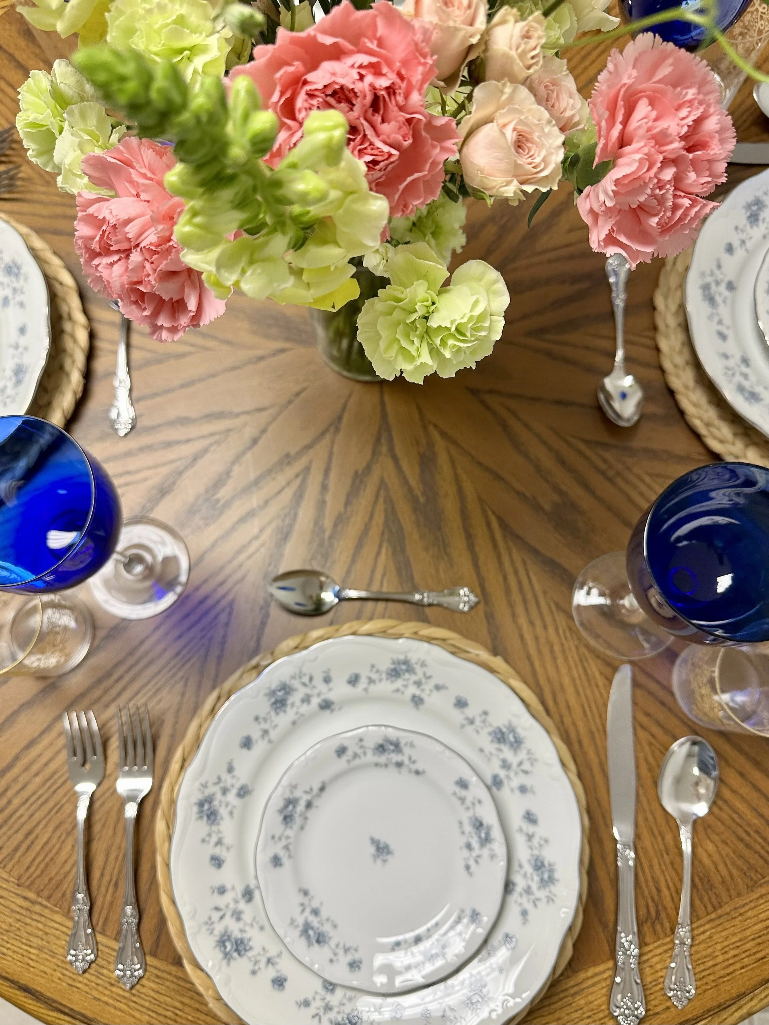 A round wooden dining table set with three place settings, each with a plate, fork, knife, and spoon, and a floral centerpiece made of pink and white carnations, green snapdragons, and white roses, with blue wine glasses and woven charger placemats.