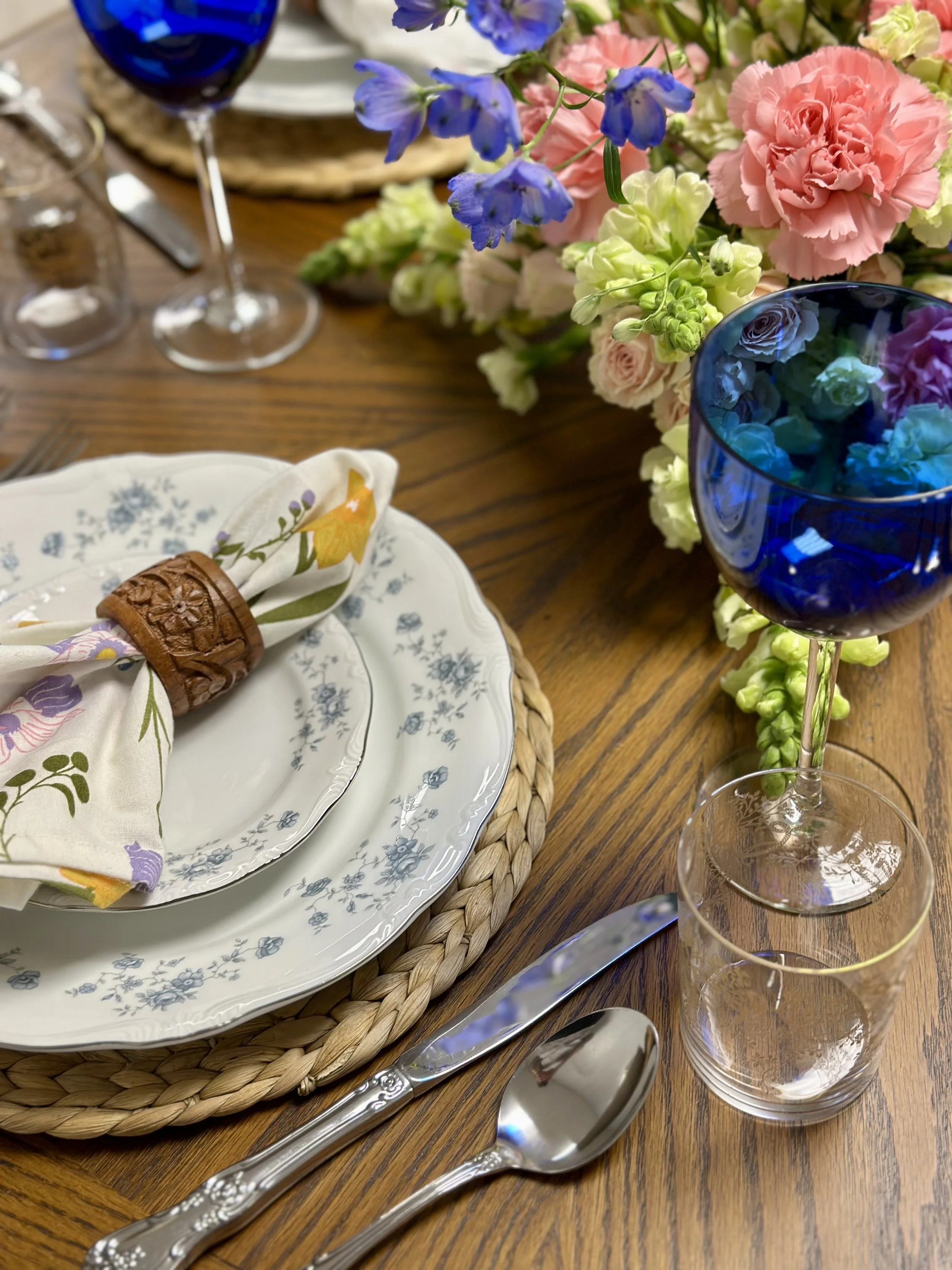 A dining table set with floral china, a floral napkin with a wooden napkin ring, two clear glasses, a blue wine glass filled with flowers, and a large colorful flower arrangement.