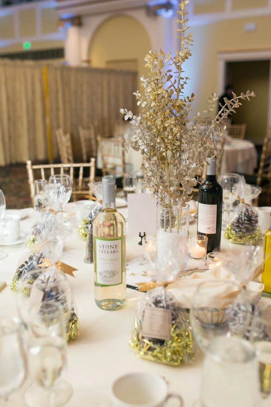 Decorated banquet table with gold and clear ornament centerpieces, wine bottles, wrapped gifts, candles, and gold chairs, set in an elegant event hall.