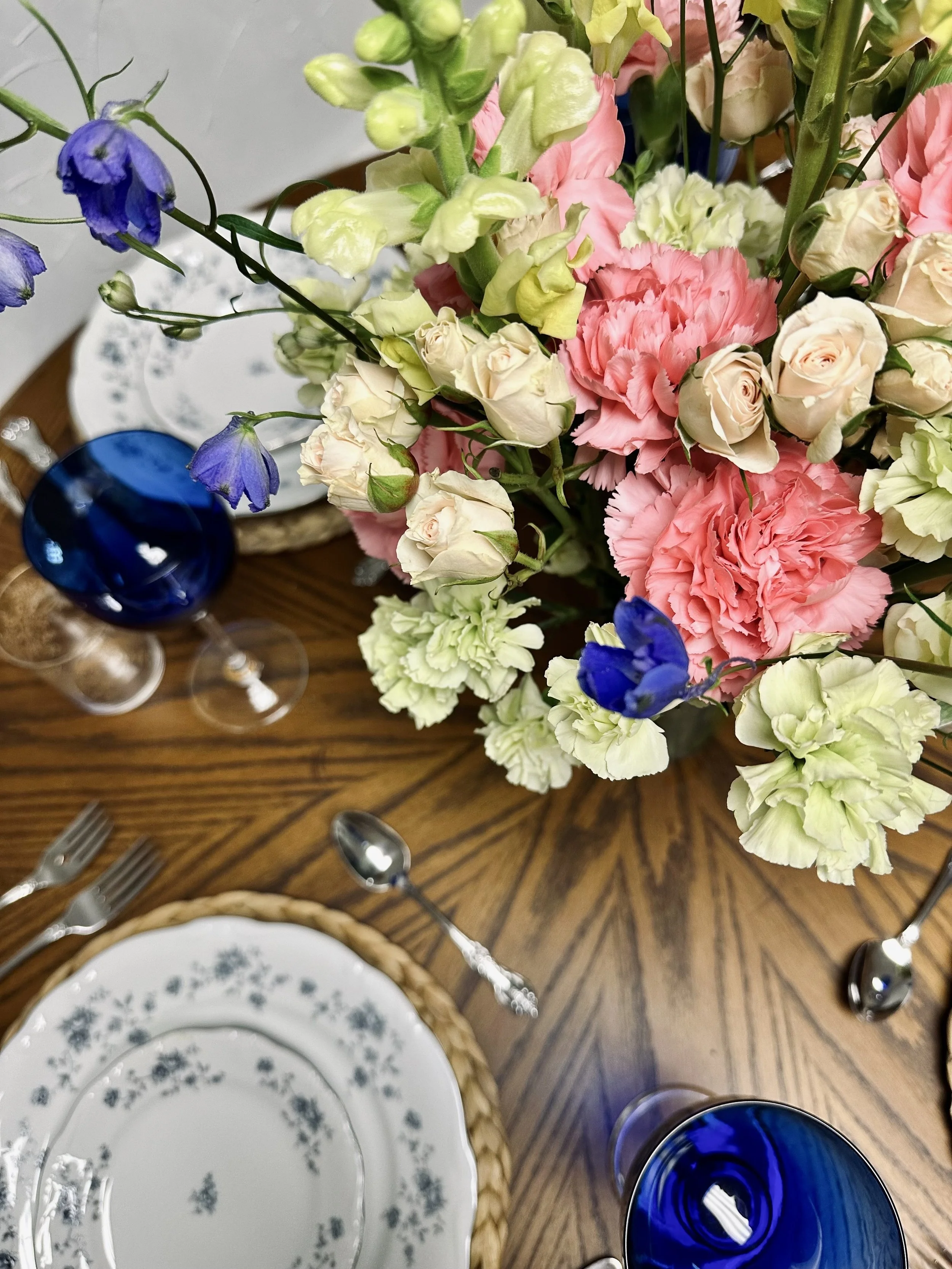 A wooden dining table set with floral centerpiece, woven charger placemats, blue drinking glasses, and white plates with blue floral patterns.