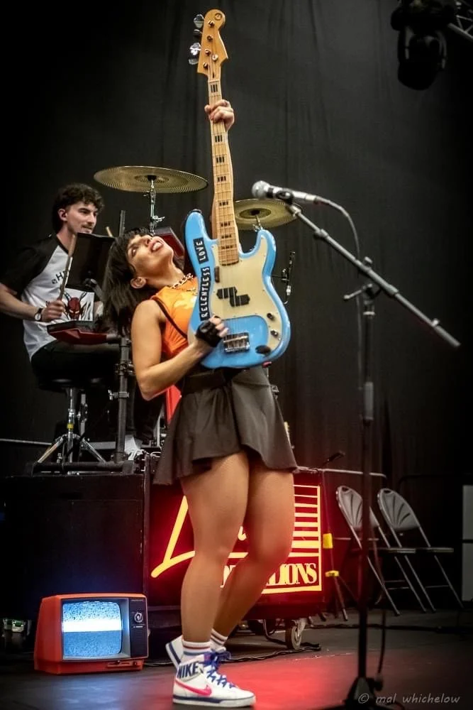 A woman on stage holding a blue electric guitar upright with her right hand, with her head tilted back and eyes closed, wearing an orange top, a dark skirt, striped Nike sneakers, and striped socks. A man behind her is playing drums, and there is a m