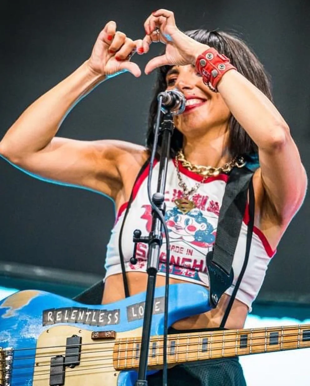 Lina with dark hair and a big smile, holding her hands in the shape of a heart above her head while playing an electric guitar. She is wearing a sleeveless top, chunky necklaces, and colorful accessories, standing in front of a microphone on a stage.