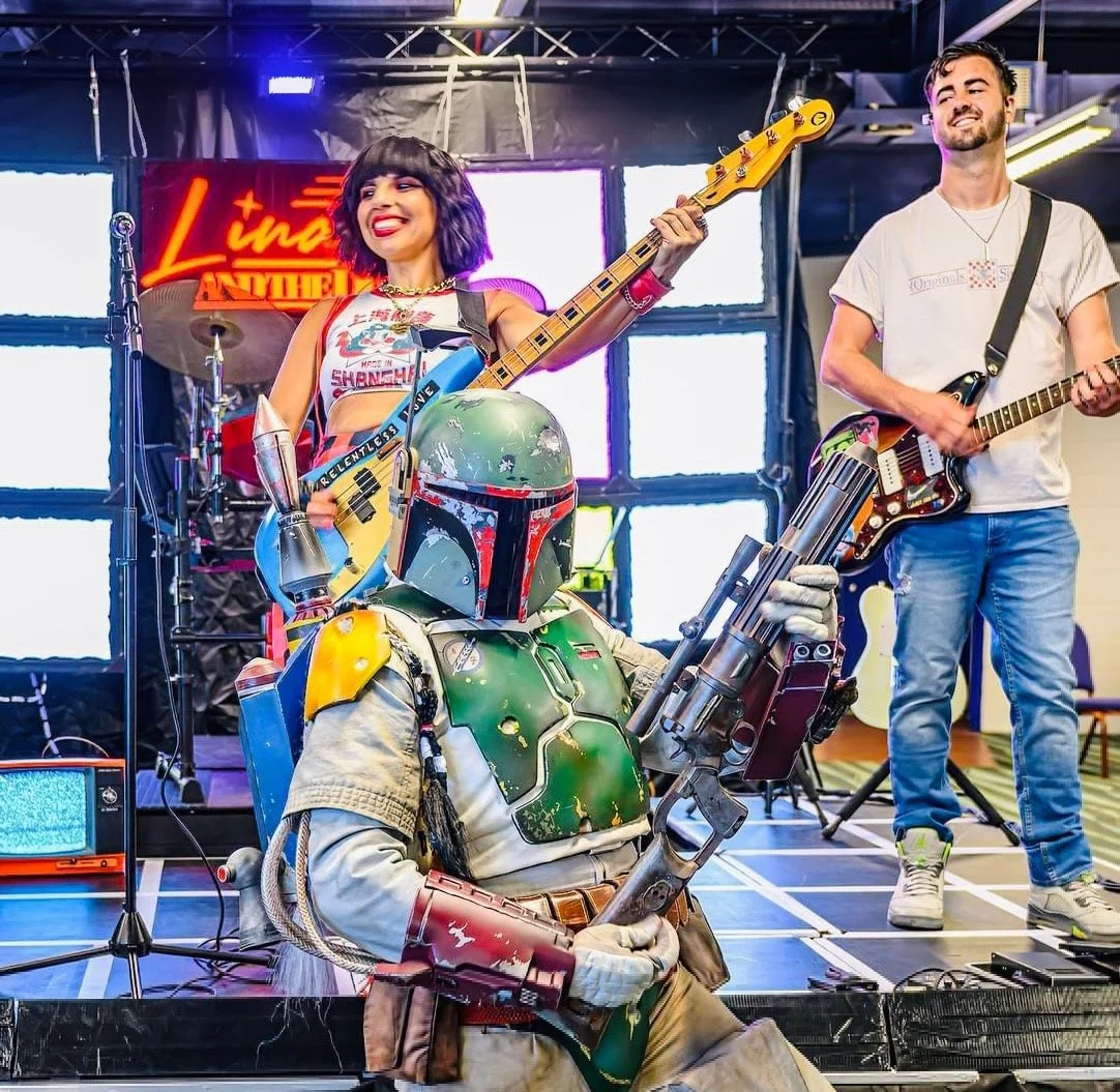 A woman with black hair and colorful makeup holding a bass guitar on stage, smiling; a man with a guitar smiling; a person dressed as Boba Fett from Star Wars in costume, holding a large weapon, seated on the stage; a neon sign with red text in the b