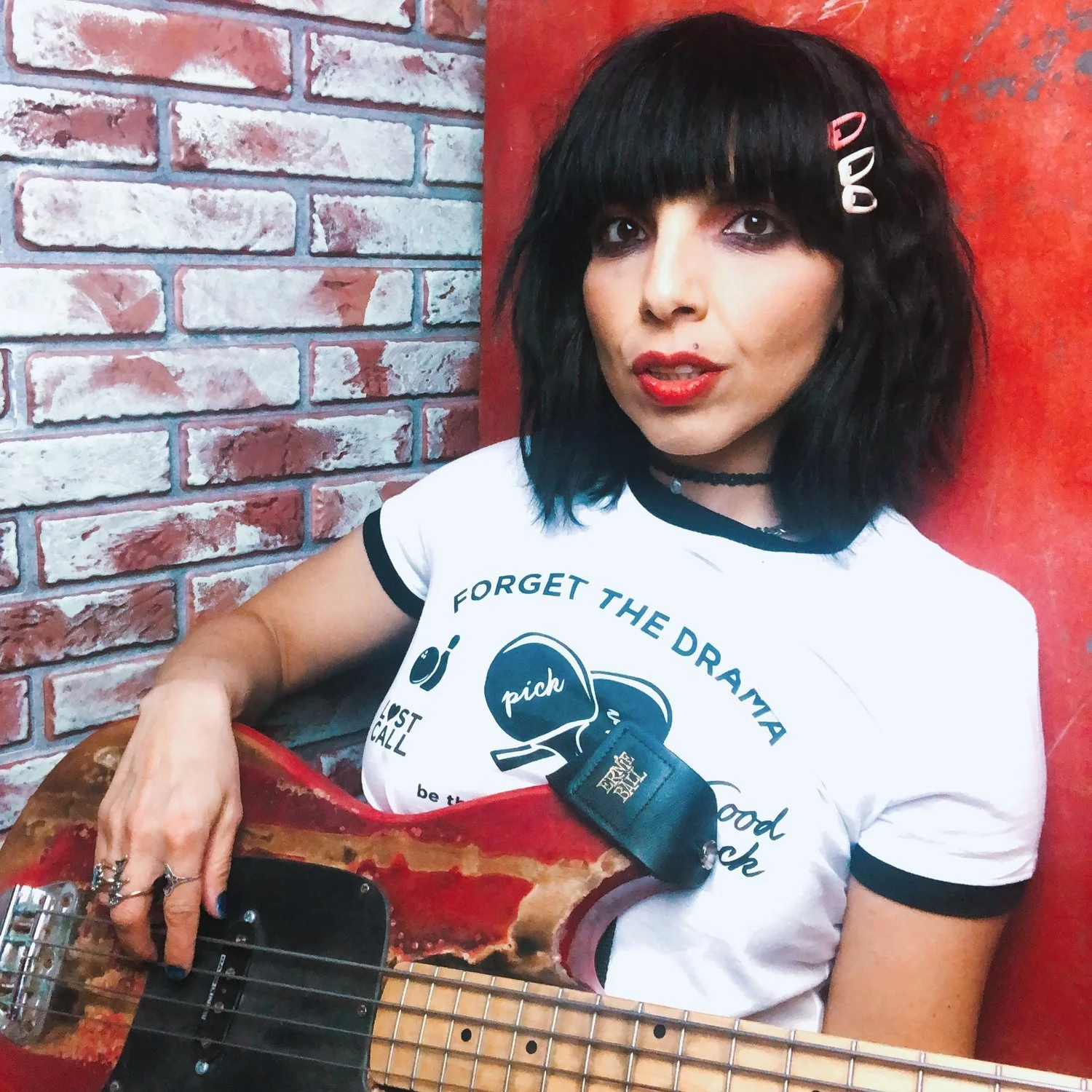 Lina Lane with black hair and bangs holding a red bass guitar, wearing a white T-shirt with the slogan 'Forget the Drama' and accessories including hair clips and rings, standing against a brick wall and red background