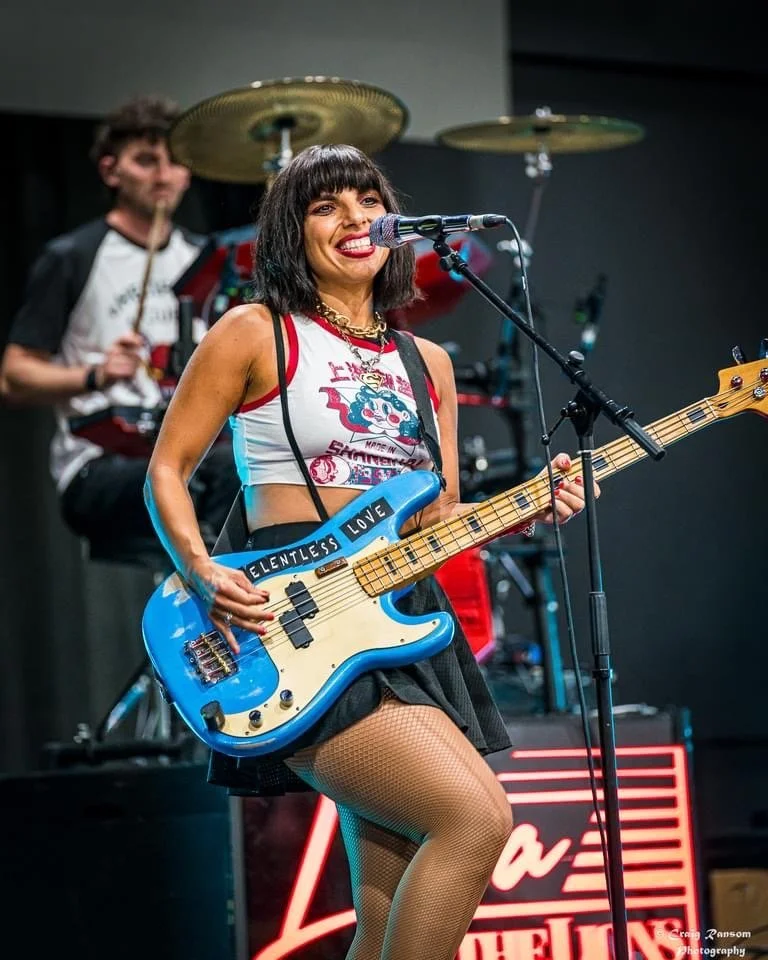 A woman with black hair and bangs playing a blue electric bass guitar, smiling into a microphone. She wears a sleeveless top with a graphic design, a black skirt, fishnet stockings, and has a sticker that says "Relentless Love" on her guitar. A drumm
