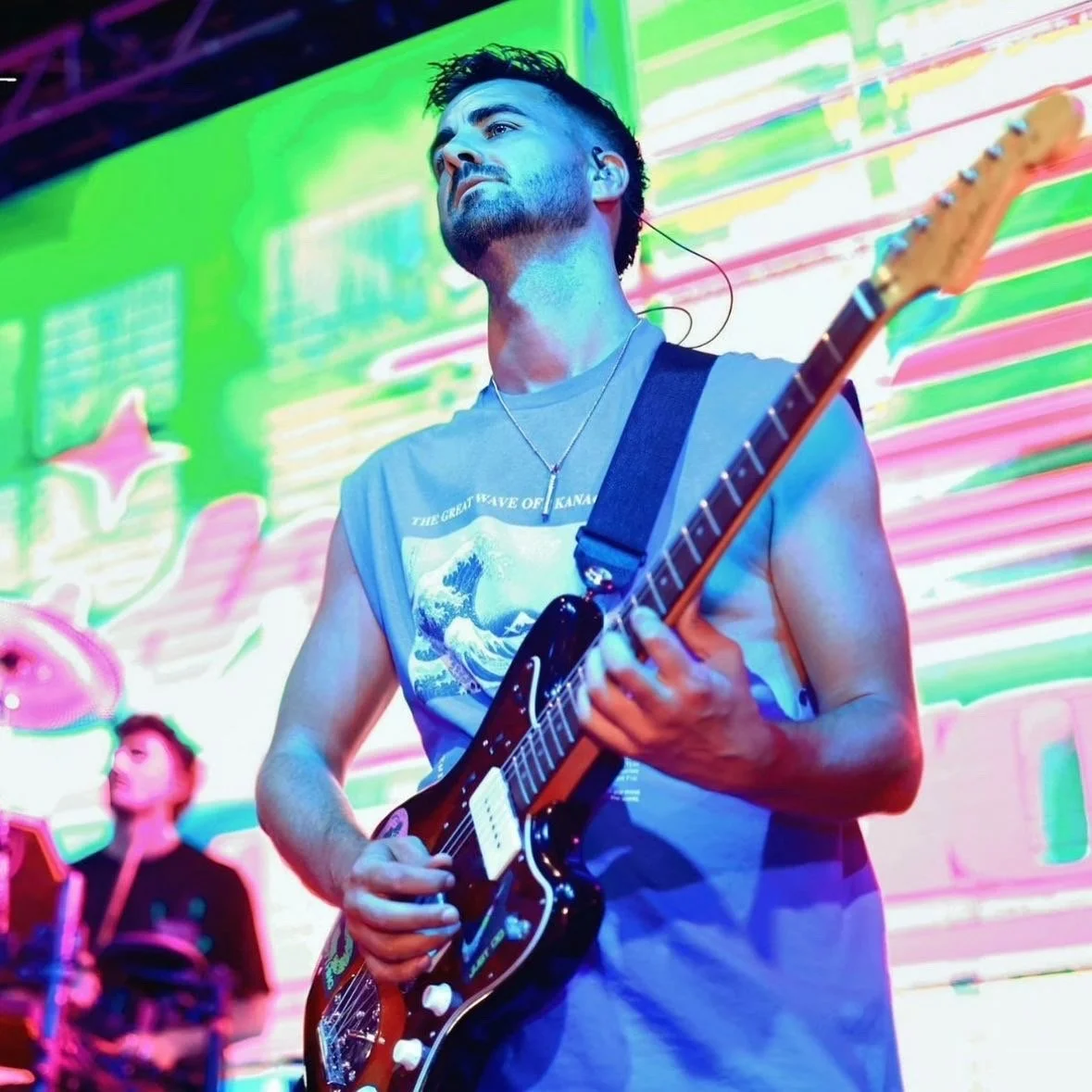 Lee Feltham guitarist performing on stage with colorful, neon background lights. He is wearing a sleeveless T-shirt, has a short beard, and is playing an electric guitar.