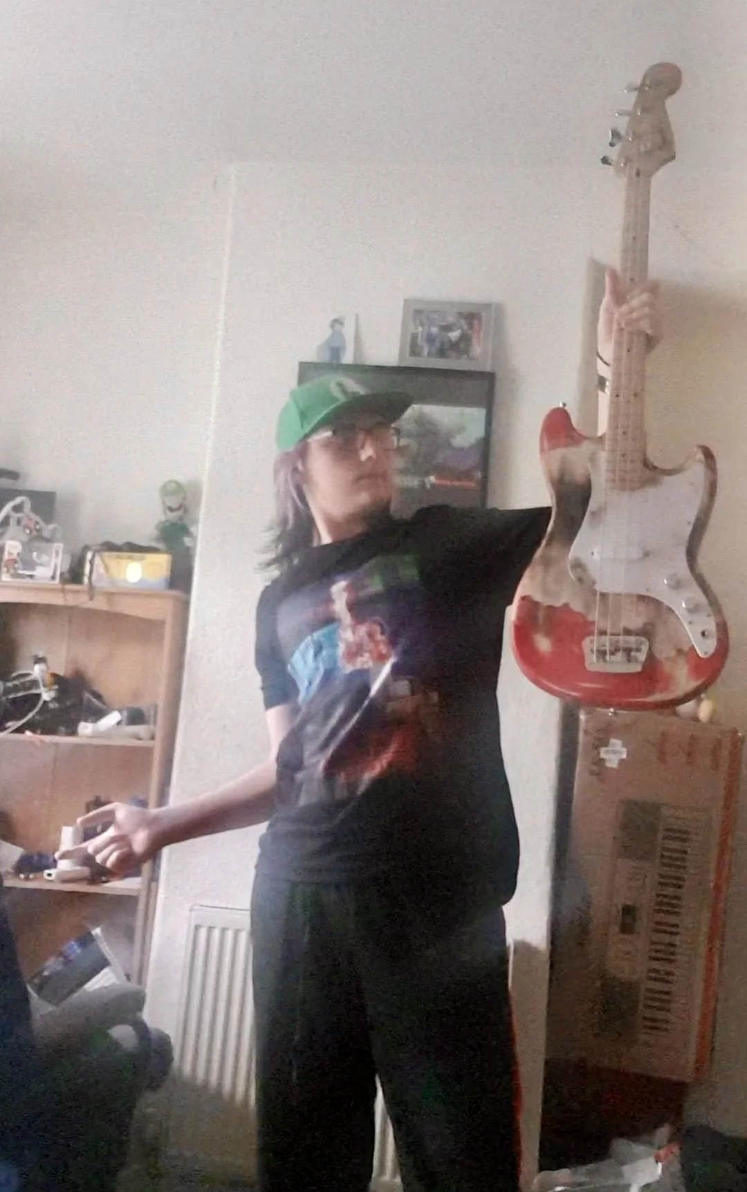 A person in a black shirt and green cap is holding an Burnie guitar and posing in a room with shelves, boxes, and photos on the wall.