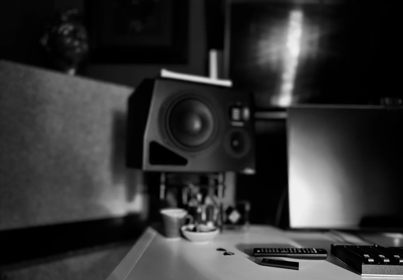 Close-up of a speaker on a desk with some electronic equipment and a computer monitor in the background, in black and white.