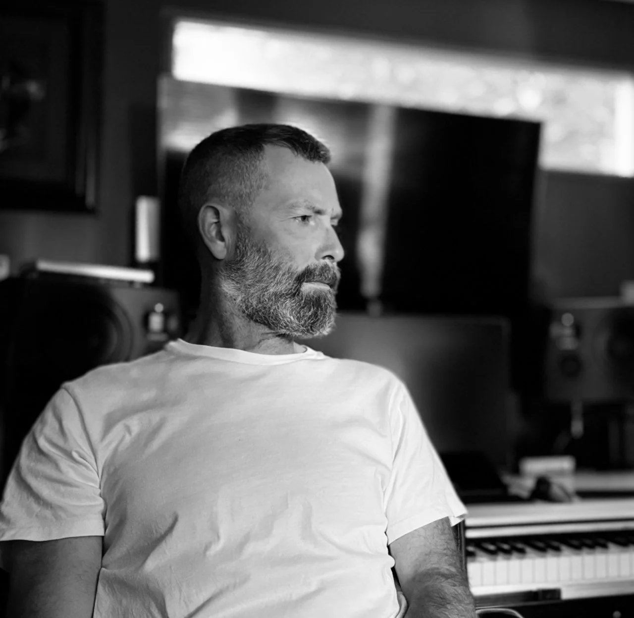 A black and white photo of a man with short hair and a beard, sitting in a studio with music equipment in the background, looking to the right.