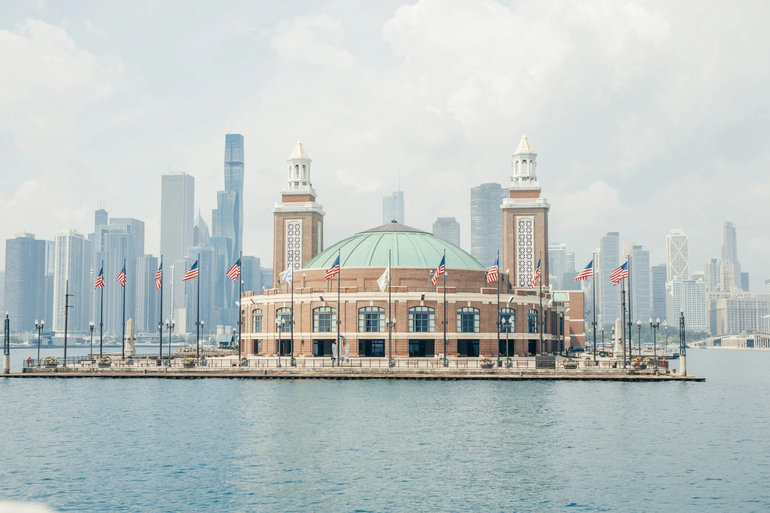 A historic building with two towers and a green-domed roof, adorned with American flags, set against a modern city skyline with tall skyscrapers in the background, located near a waterfront.