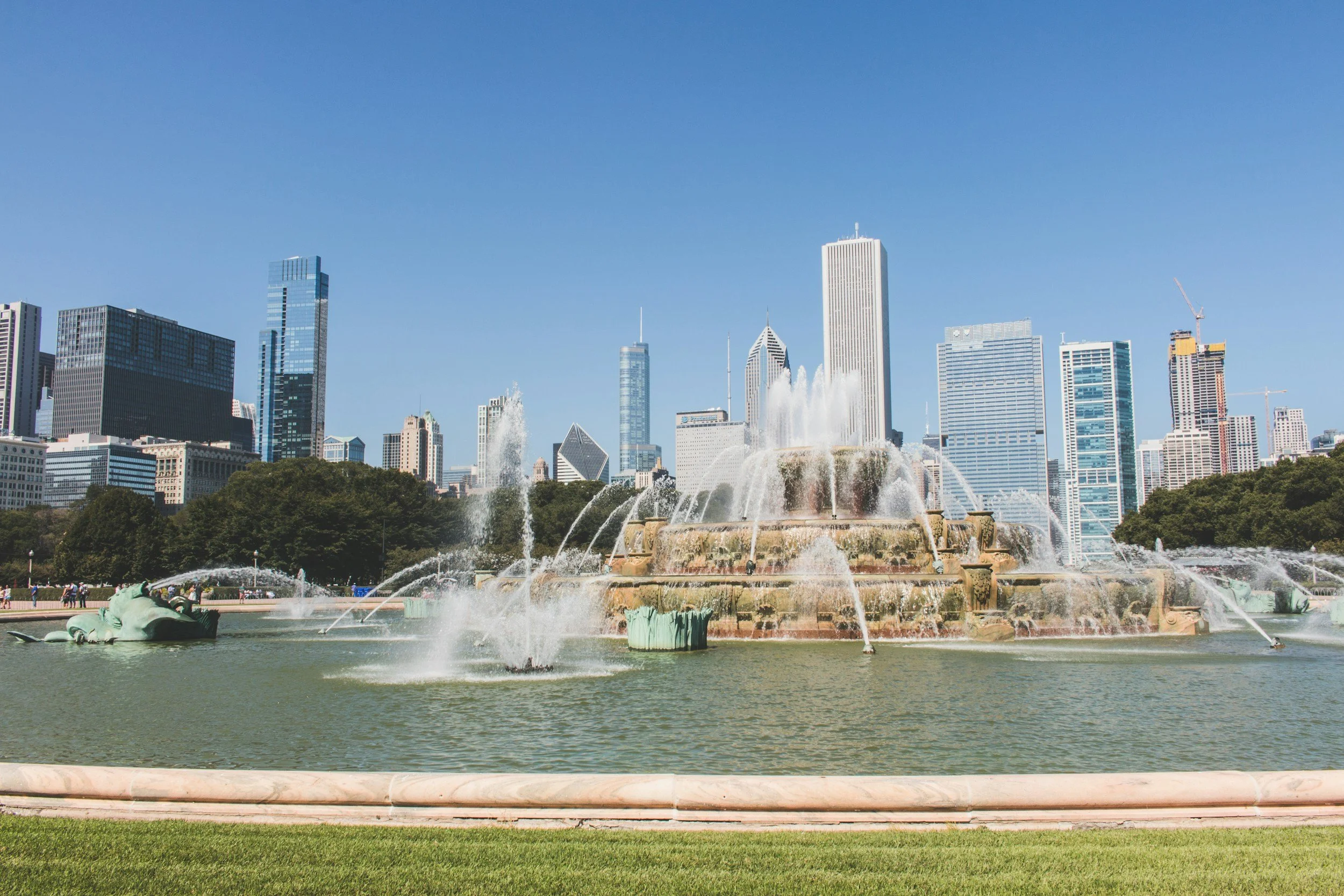 Chicago skyline with tall skyscrapers including the 875 North Michigan Avenue building, and the Buckingham Fountain in Grant Park with water spraying from its ornate bronze statues, under a clear blue sky.