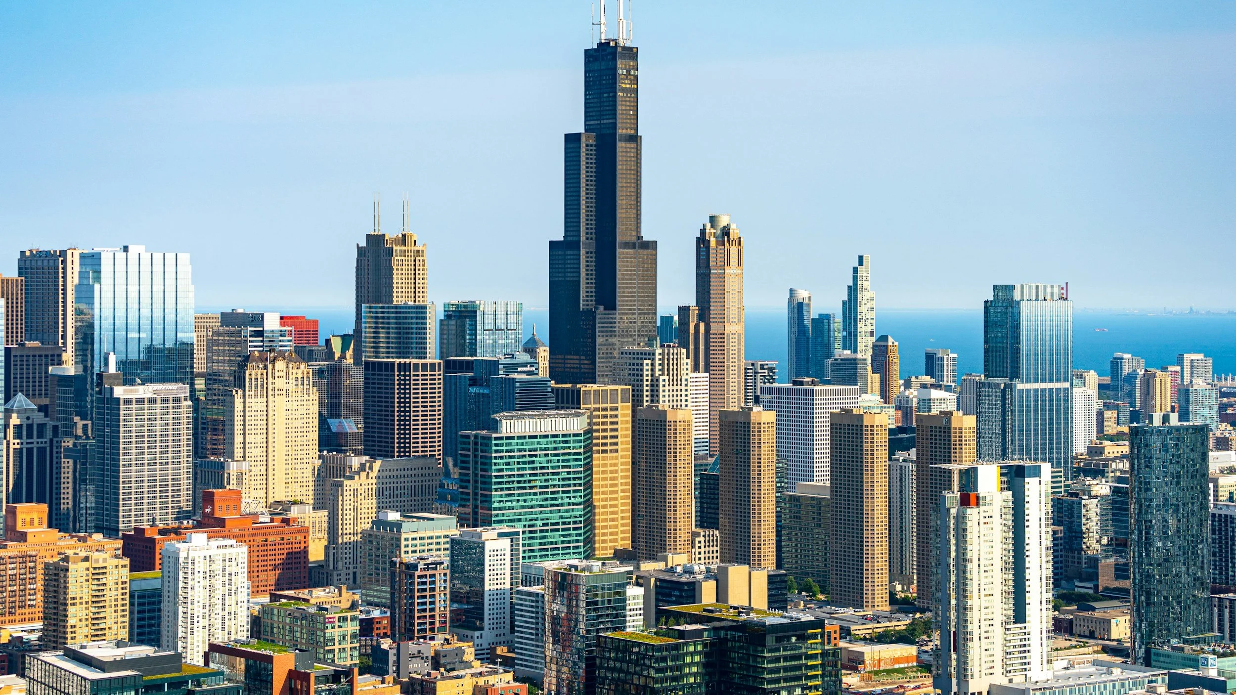 Skyline view of Chicago with numerous tall modern skyscrapers, including the Willis Tower, against a clear blue sky and a hint of Lake Michigan in the background.