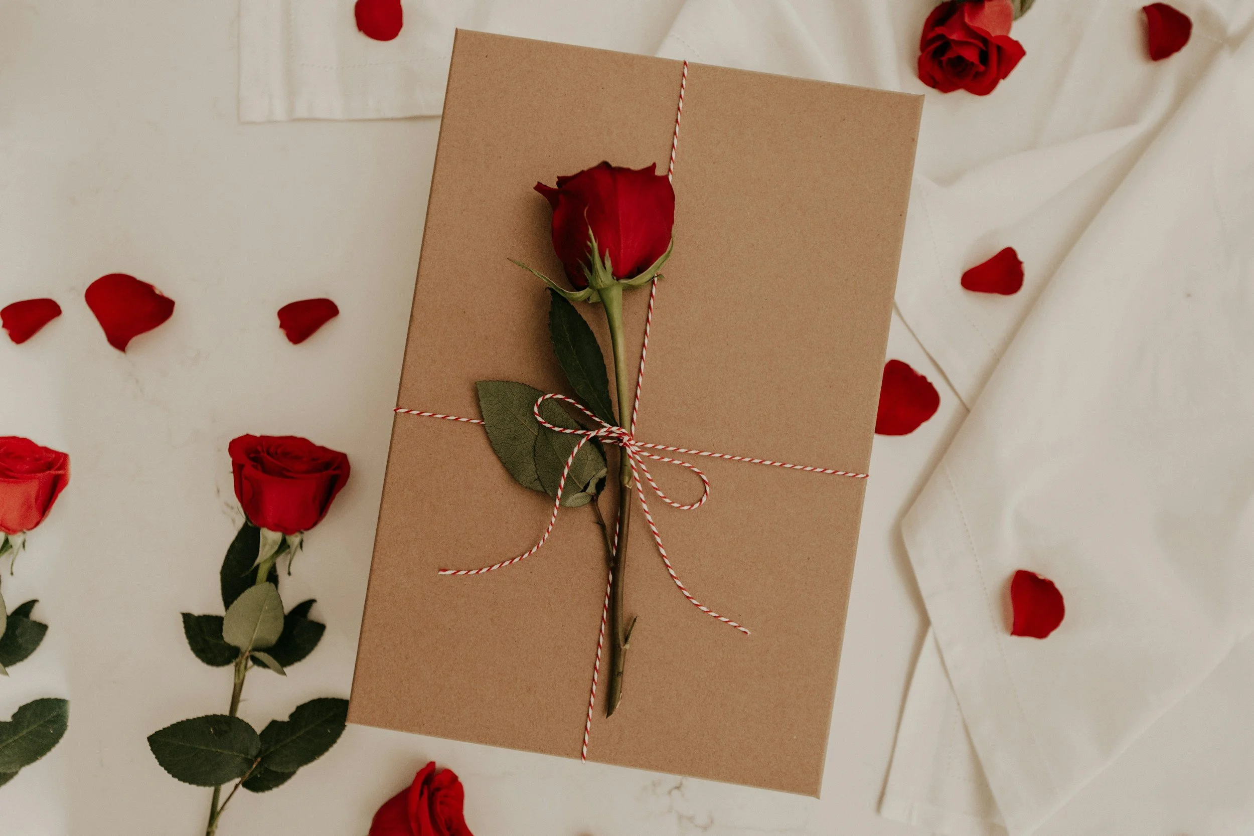 A gift wrapped in plain brown paper, tied with red and white string, with a single red rose attached to the top. The background has scattered red rose petals and green rose stems on a white surface.